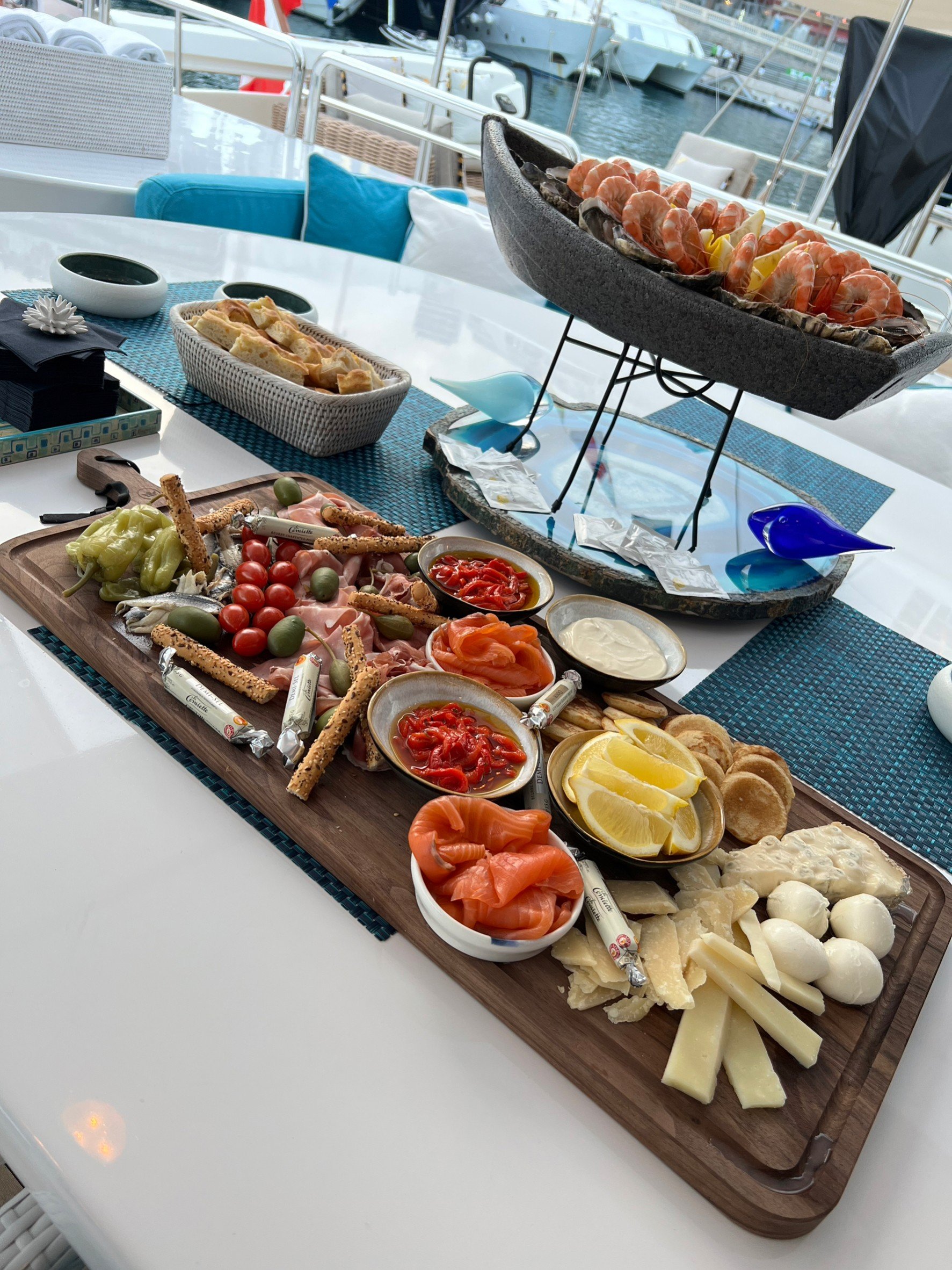 A yacht's table set with a seafood platter, cheese, crackers, and various appetizers, with boats docked in the marina in the background.
