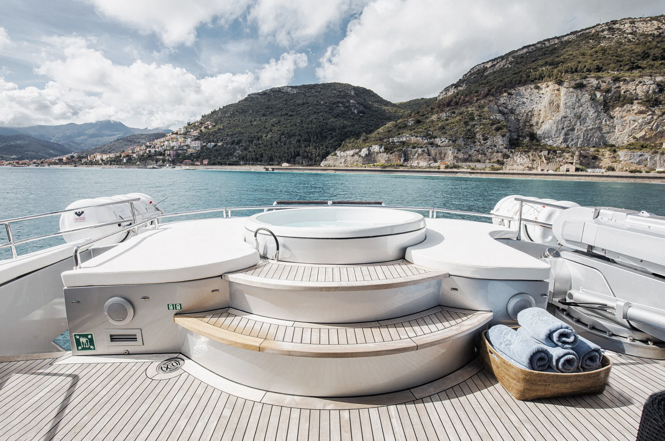 View of the deck of a luxury yacht with a hot tub, soft blue towels in a basket, and scenic mountains and water in the background.