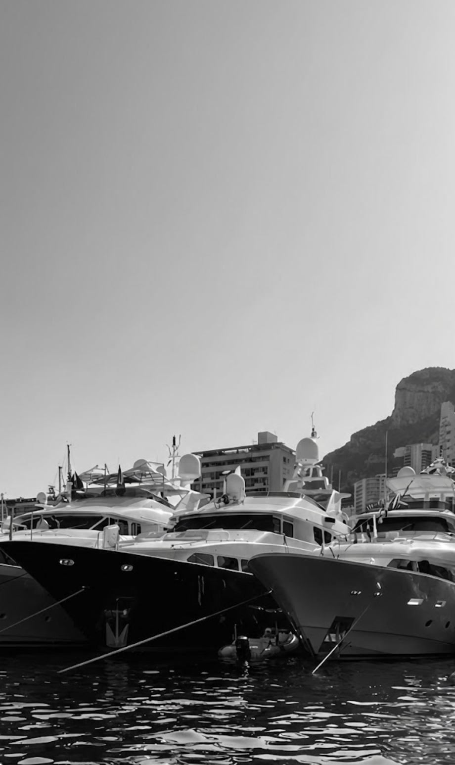 Several luxury yachts docked at a marina with buildings and a mountain in the background, black and white photograph.