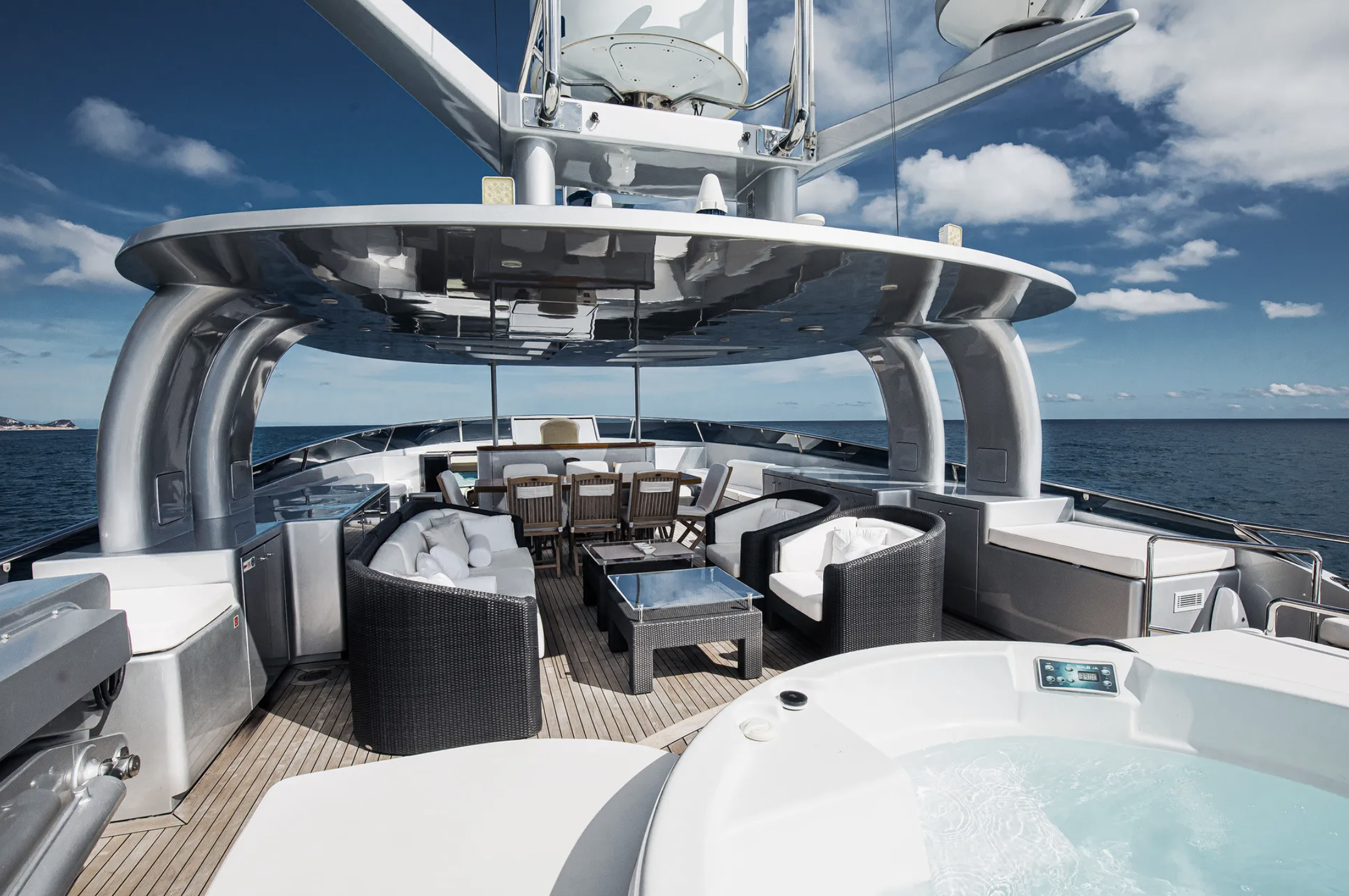 Luxury yacht deck with seating, dining area, and hot tub, sails on the ocean under a partly cloudy sky.