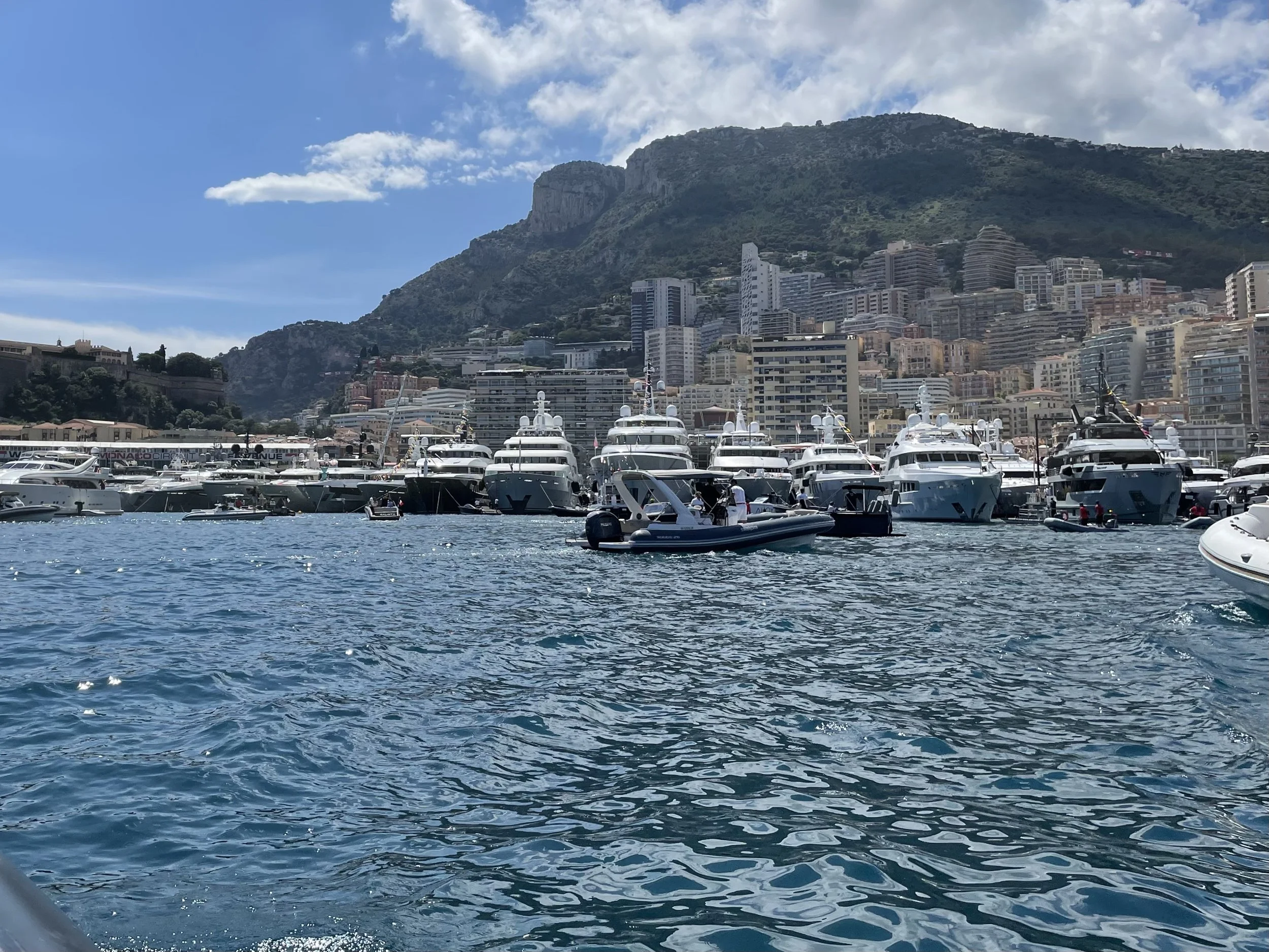 Yachts and boats docked at a marina with a city and mountain backdrop under a partly cloudy sky.