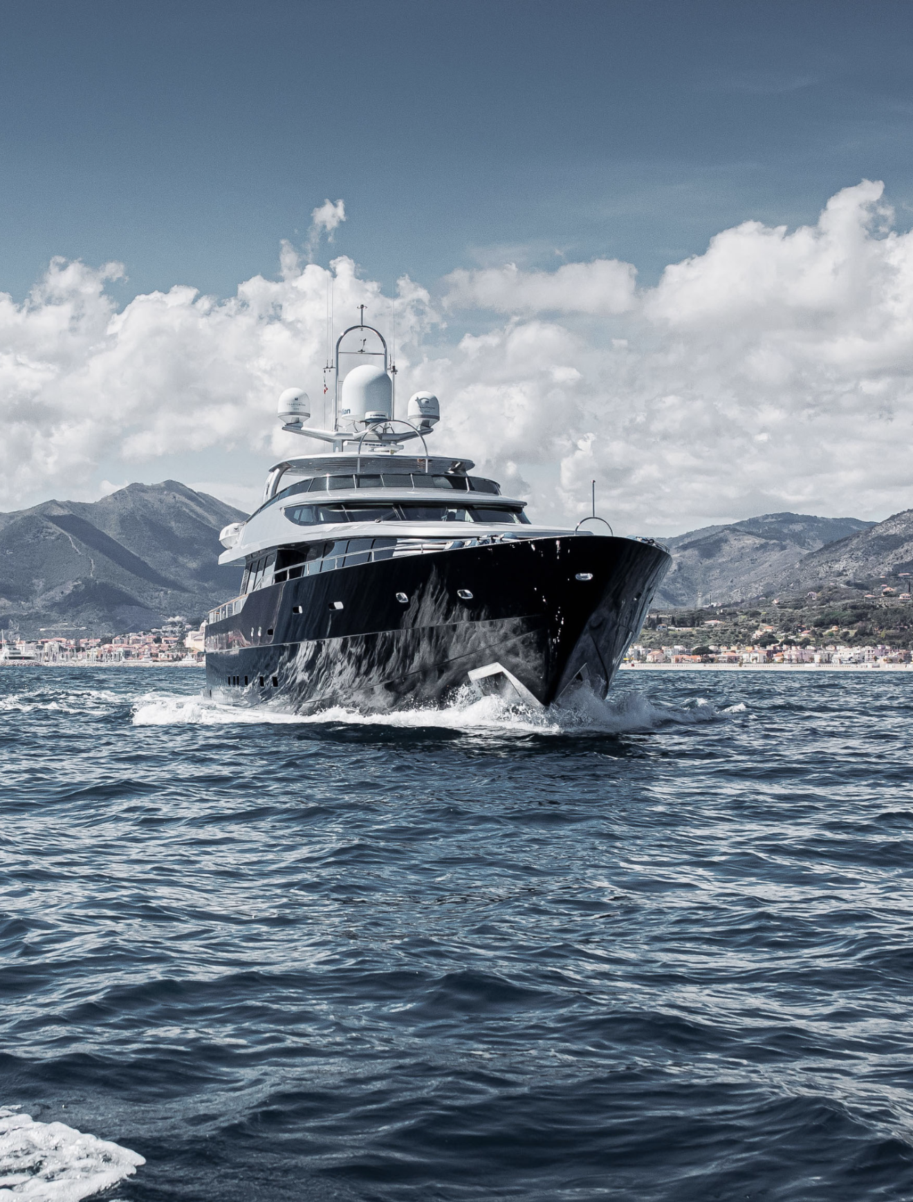 A large black yacht sailing on the water with a coastal town and mountains in the background, under a partly cloudy sky.