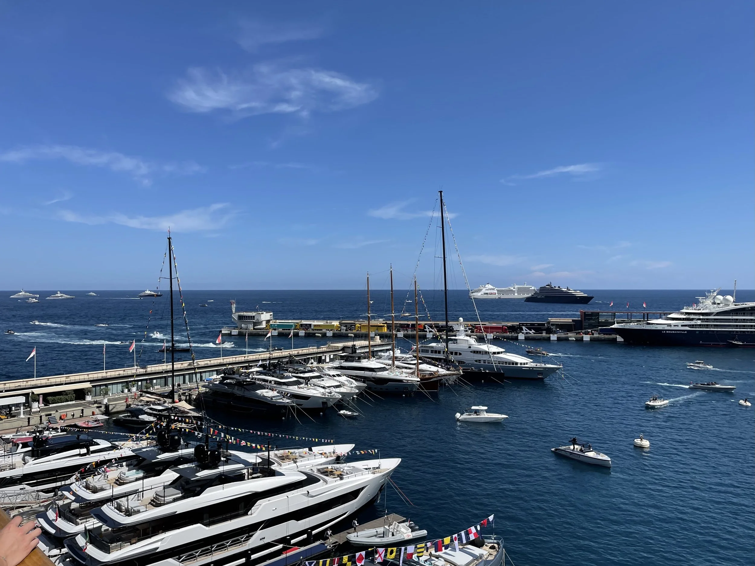 View of a marina filled with luxury yachts and boats, with a clear blue sky and calm sea in the background.