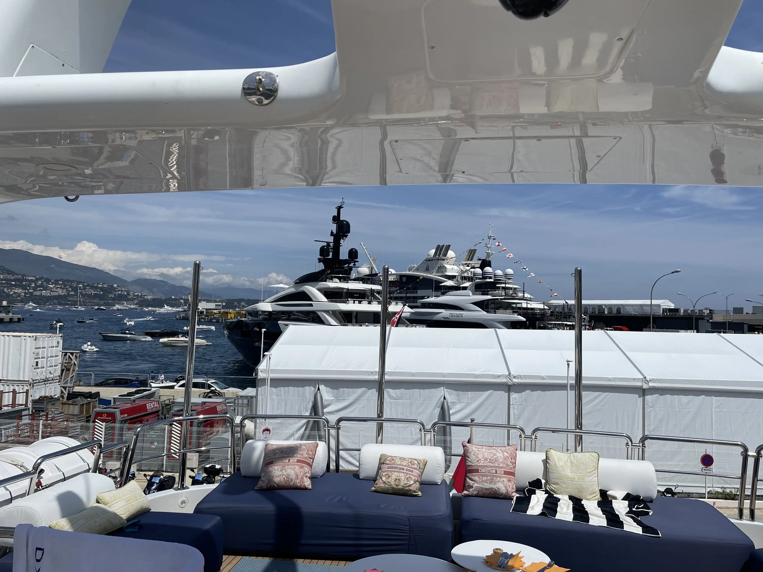 View of a luxury yacht docked at a marina with other boats in the water, a mountain range in the background, and chairs and pillows on the yacht's deck in the foreground.