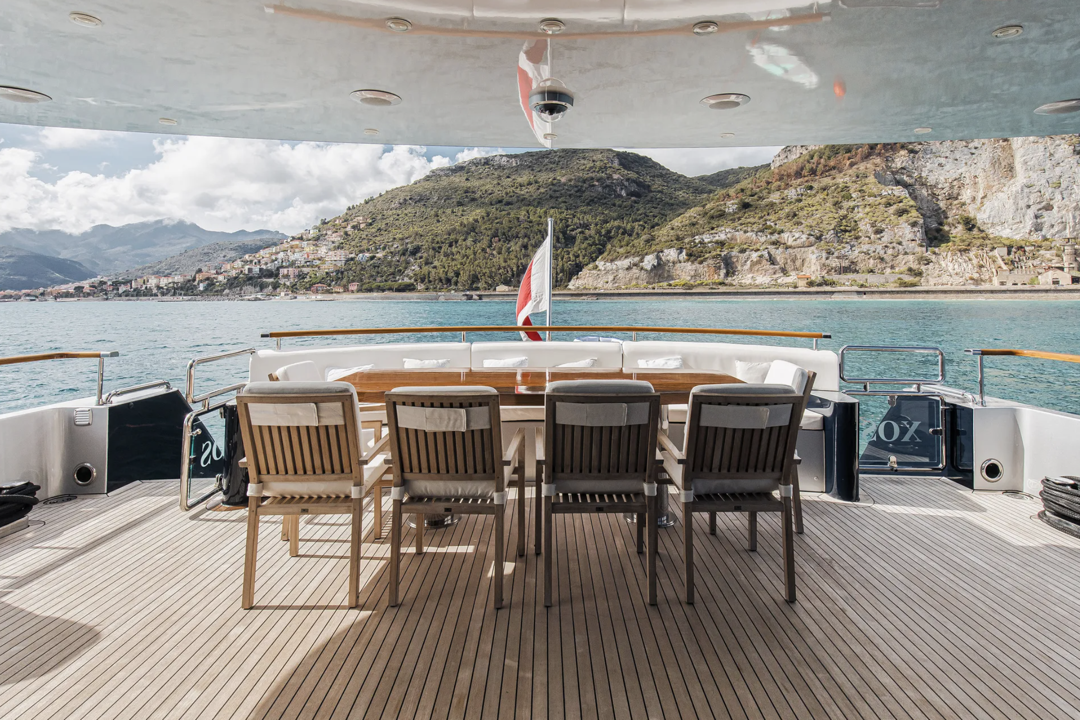 View from the deck of a yacht with a wooden table and chairs, overlooking a body of water with hills and mountains in the distance.
