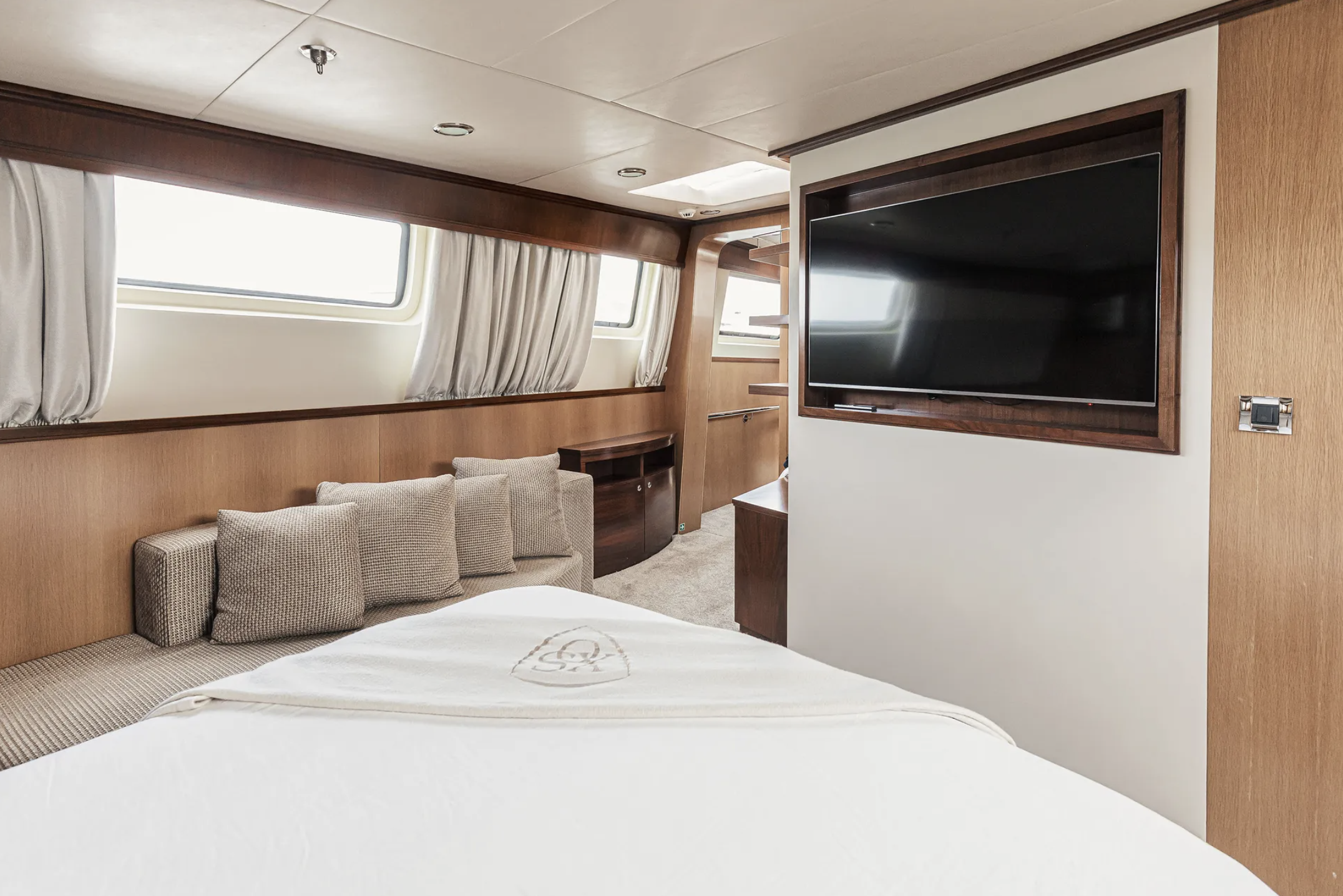 A cruise ship cabin with a bed, a sofa with pillows, a large window with curtains, and a wall-mounted TV.