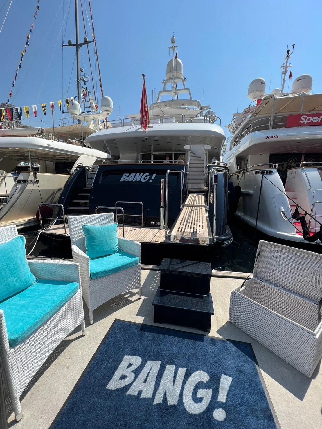 Luxury yachts docked at a marina with outdoor furniture including wicker chairs with teal cushions, a black table, and a blue rug with the word 'BANG!' in white, on a sunny day with clear blue sky.