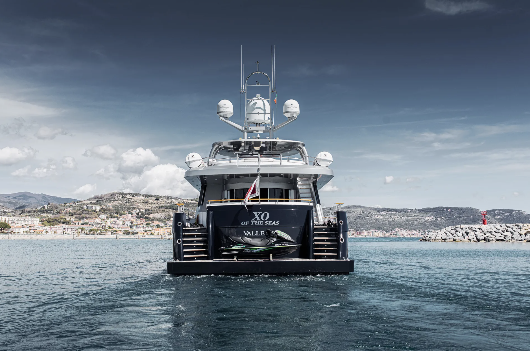 Large yacht named 'XO of the Seas' docked in harbor with a jet ski on the back. The background shows a coastal town, hills, and a lighthouse on a breakwater. The sky is partly cloudy.