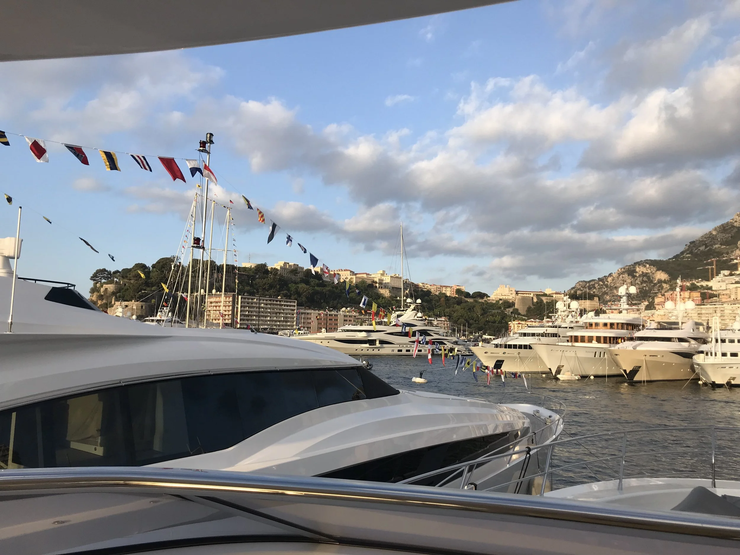 Yachts docked at a marina with colorful flags hanging from a line overhead, hillside buildings in the background, and partly cloudy sky.