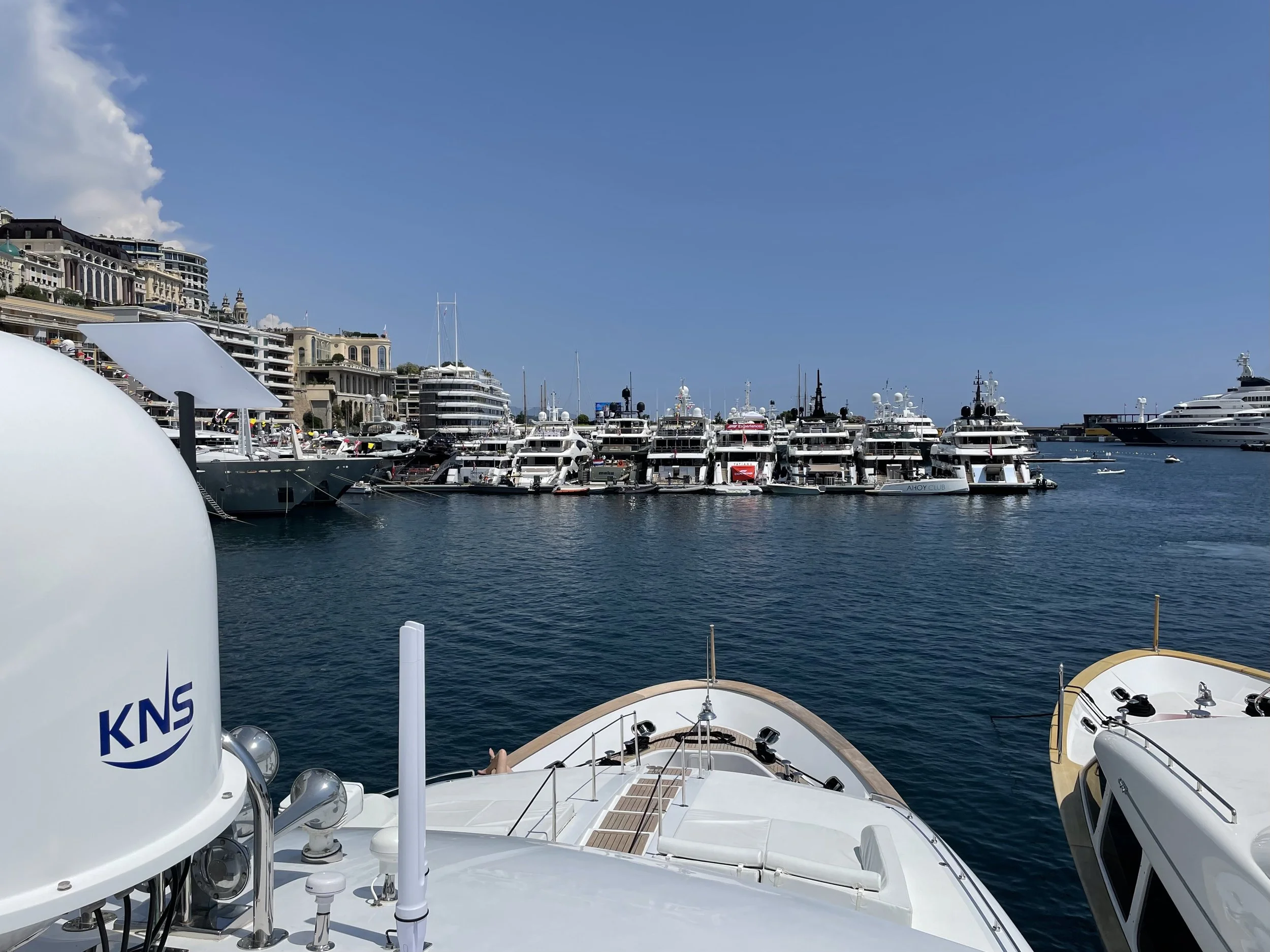 View from a luxury yacht looking over a marina filled with yachts and boats, with a cityscape and high-end buildings in the background under a clear blue sky.