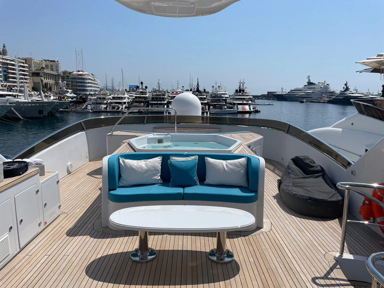View from the deck of a yacht showing a seating area with a blue cushioned sofa, a white oval table, and a hot tub, with numerous yachts and boats docked at a marina in the background.