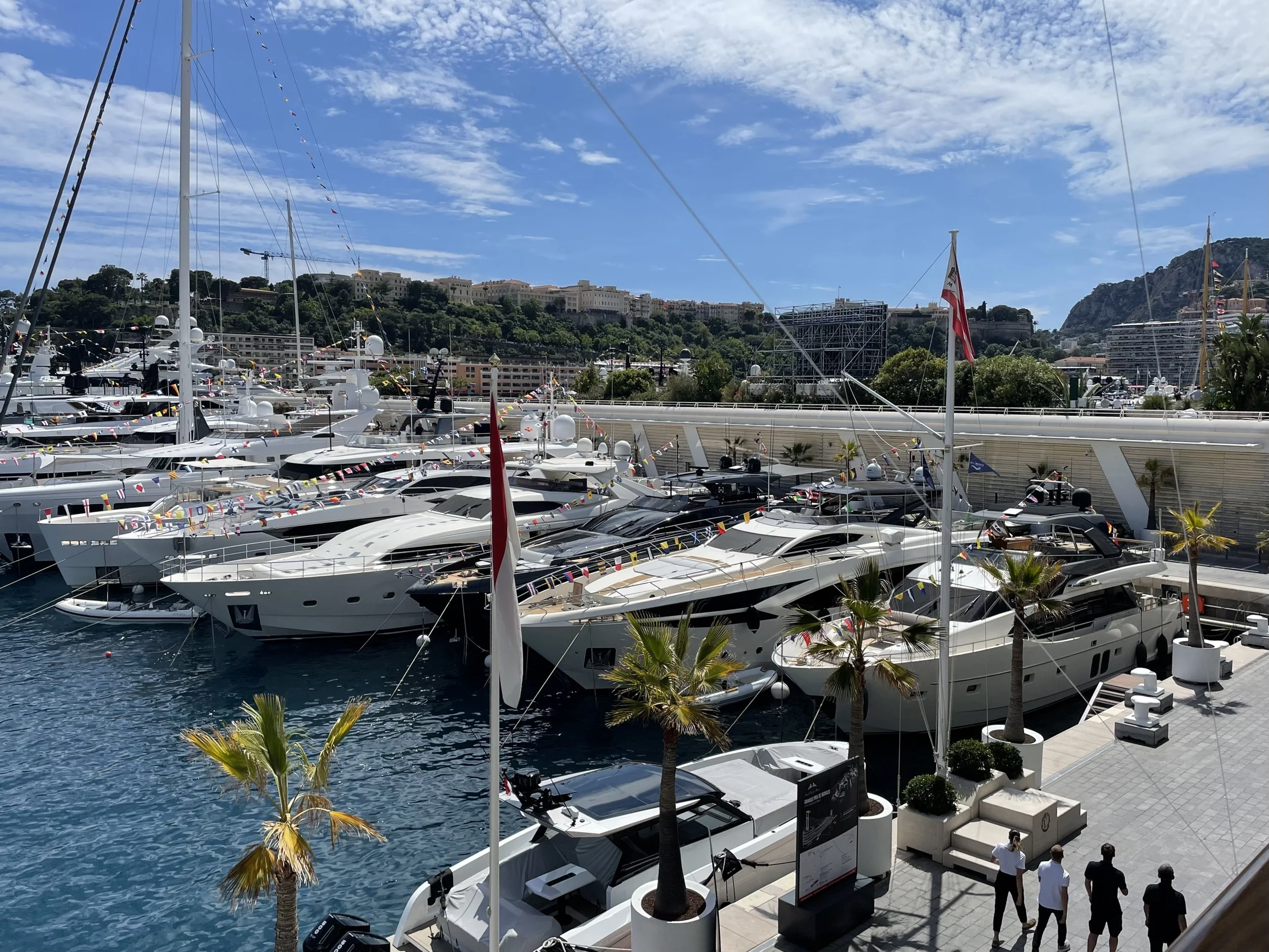 Luxurious yachts docked at a marina with palm trees, people strolling on the pier, and a hillside with buildings in the background under a partly cloudy blue sky.