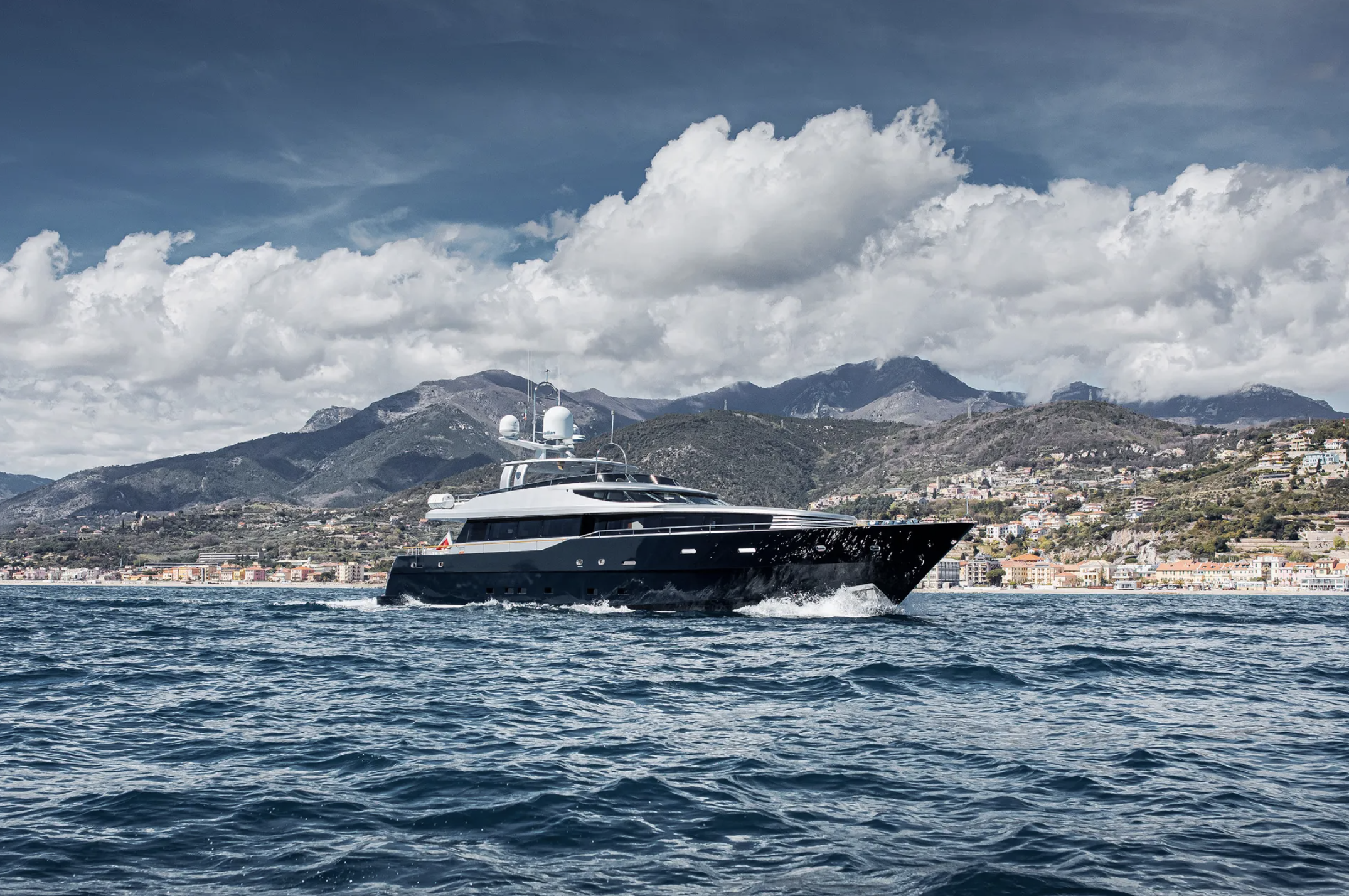 A luxury yacht sailing on the ocean with a mountainous coastline and a cloudy sky in the background.