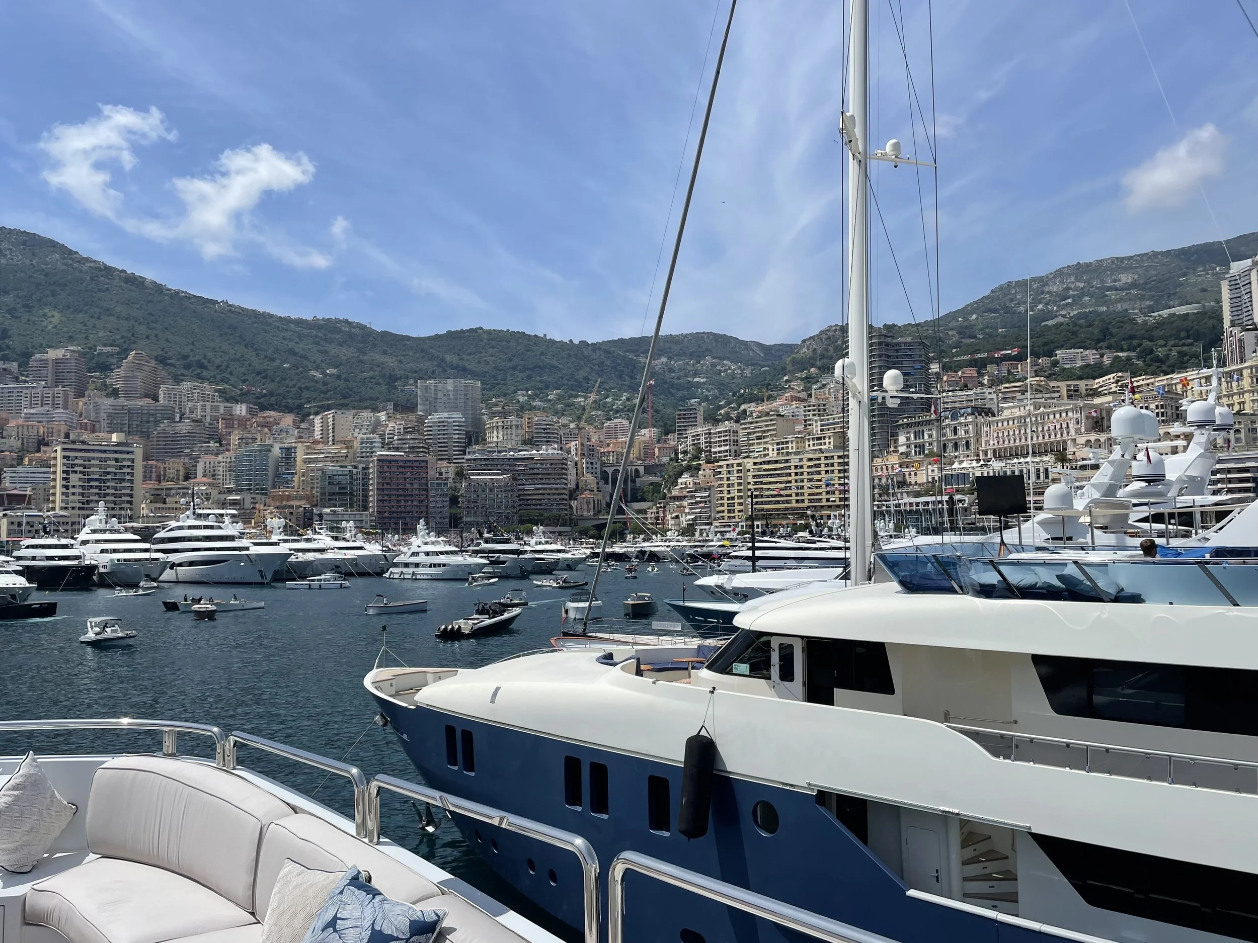View of a marina filled with yachts and boats, with a city and mountains in the background under a partly cloudy sky.