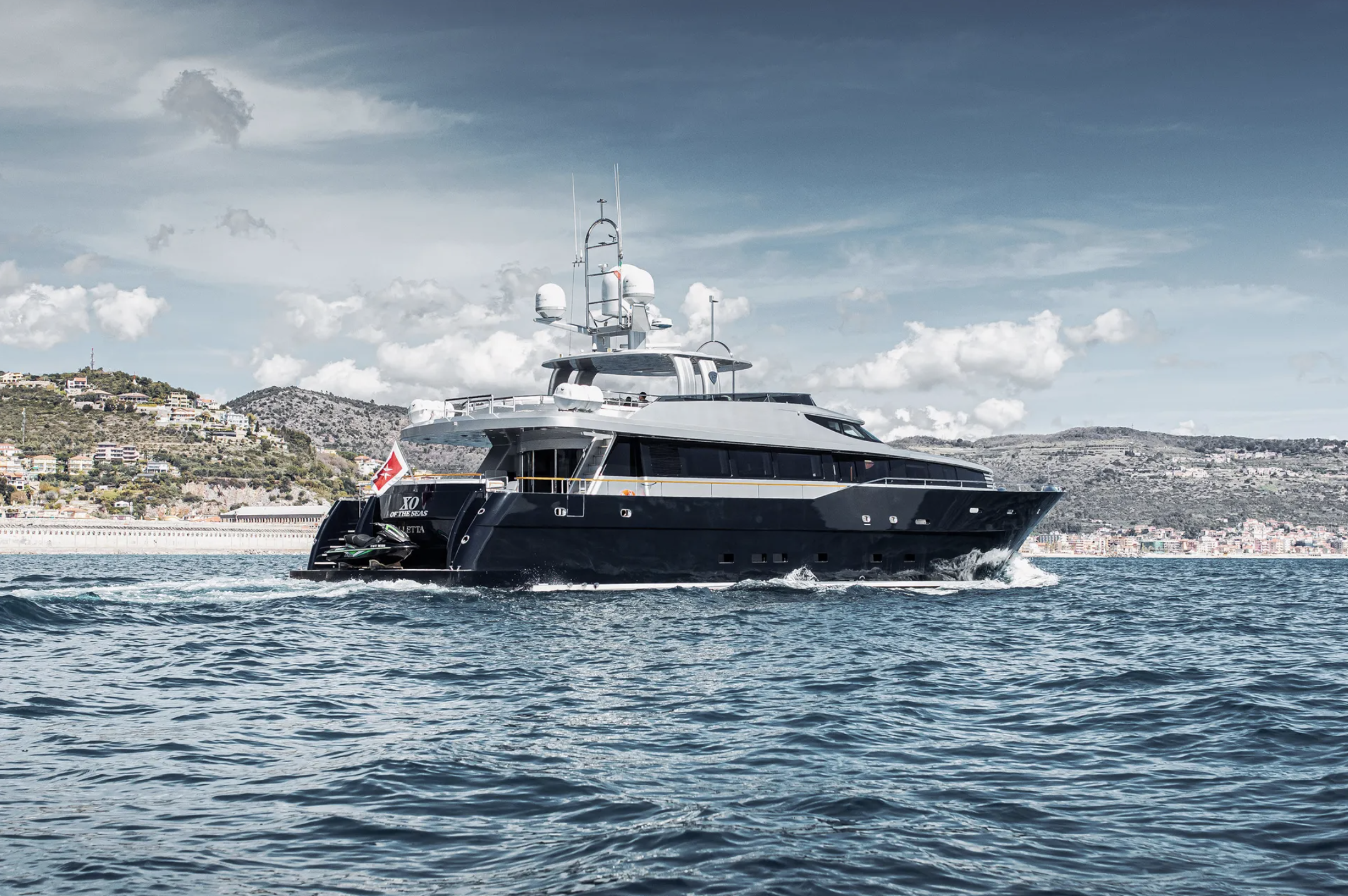A luxury yacht sailing on the ocean with a hillside city in the background and partly cloudy sky.