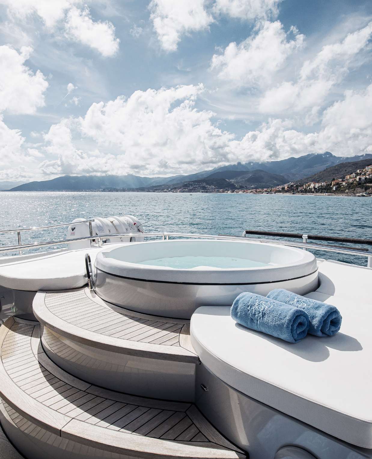View of a yacht's deck with a hot tub, two rolled blue towels, and a scenic ocean, mountains, and sky in the background.