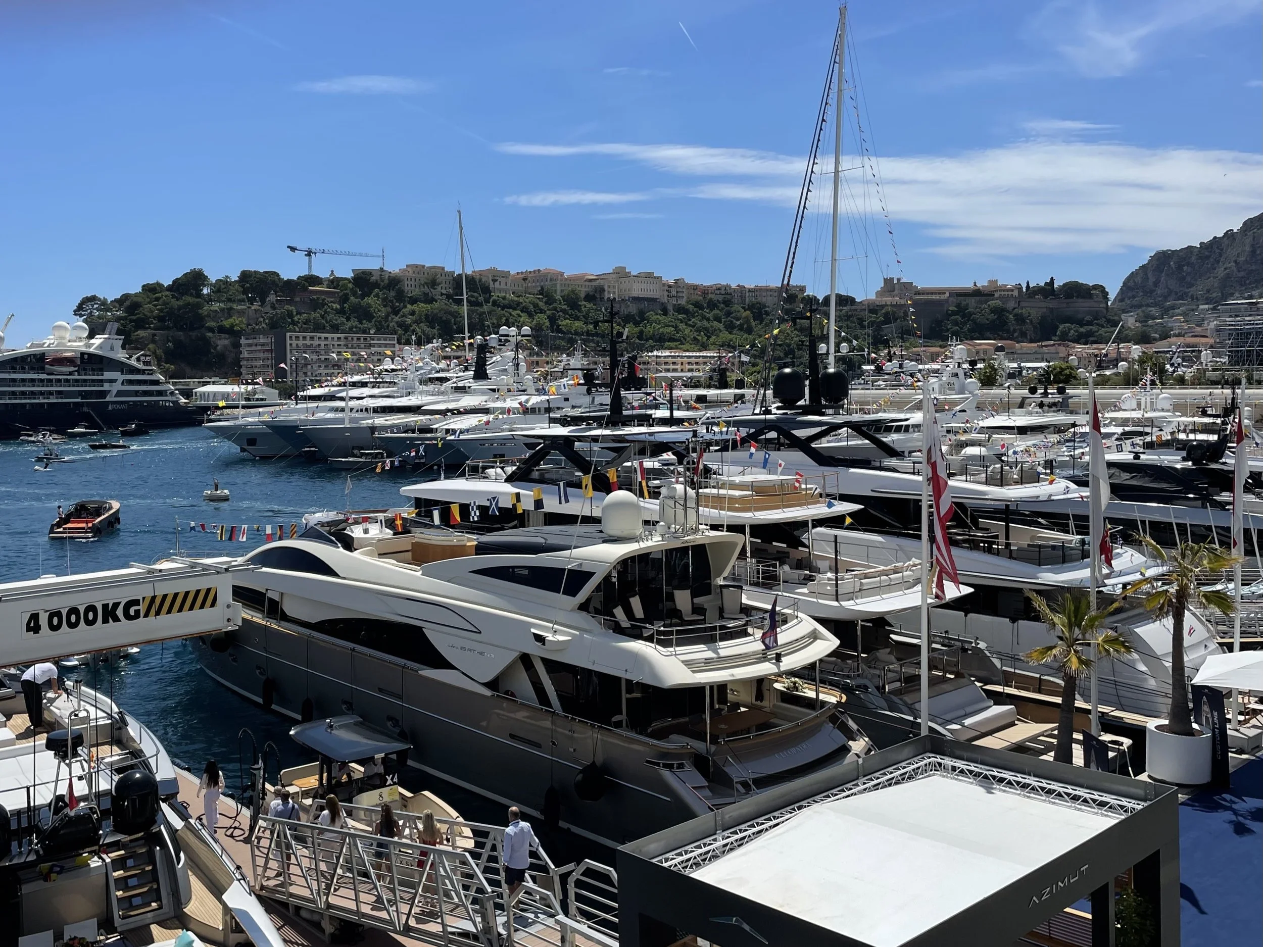 A marina filled with luxury yachts and boats docked, with a hillside cityscape and clear blue sky in the background.