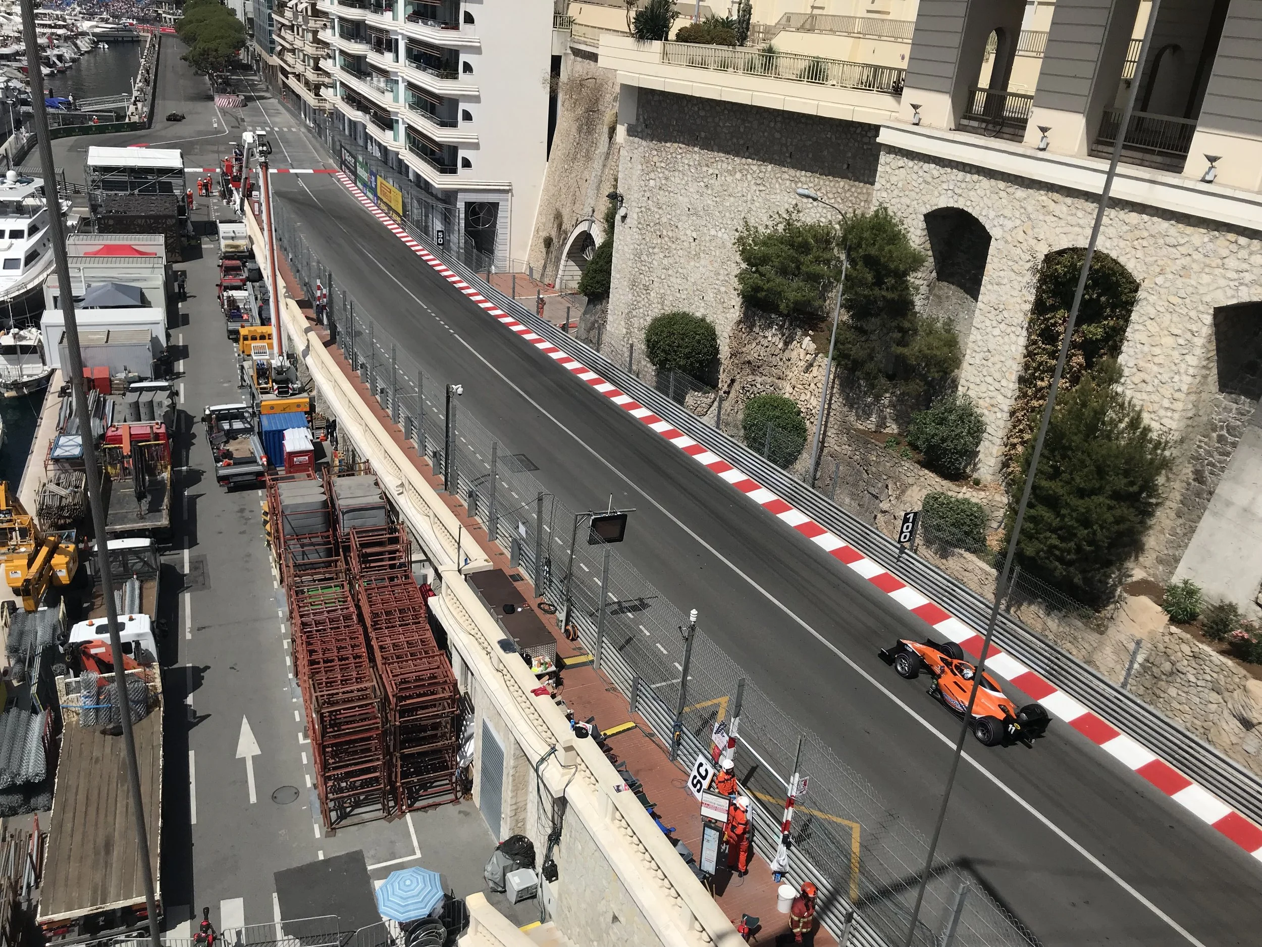 A racing car speeds down a racetrack set alongside a city street with boats docked along a waterway. The track is elevated above the street and has red and white curbs. There are safety barriers and crew members wearing orange safety vests near the t