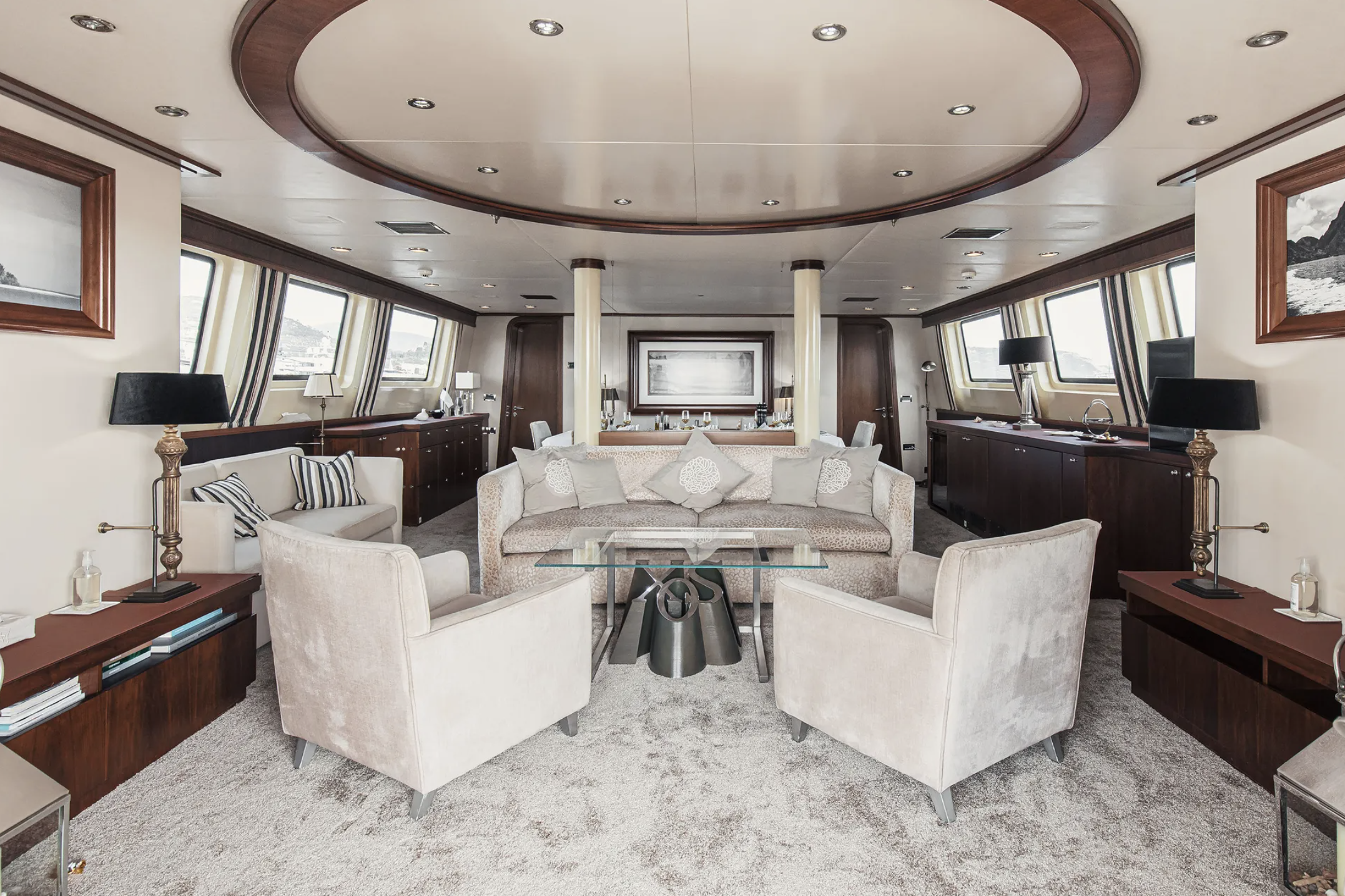 Interior of a luxurious yacht lounge with beige armchairs, a glass coffee table, dark wood sideboards, framed black-and-white photographs, and large windows with striped curtains.