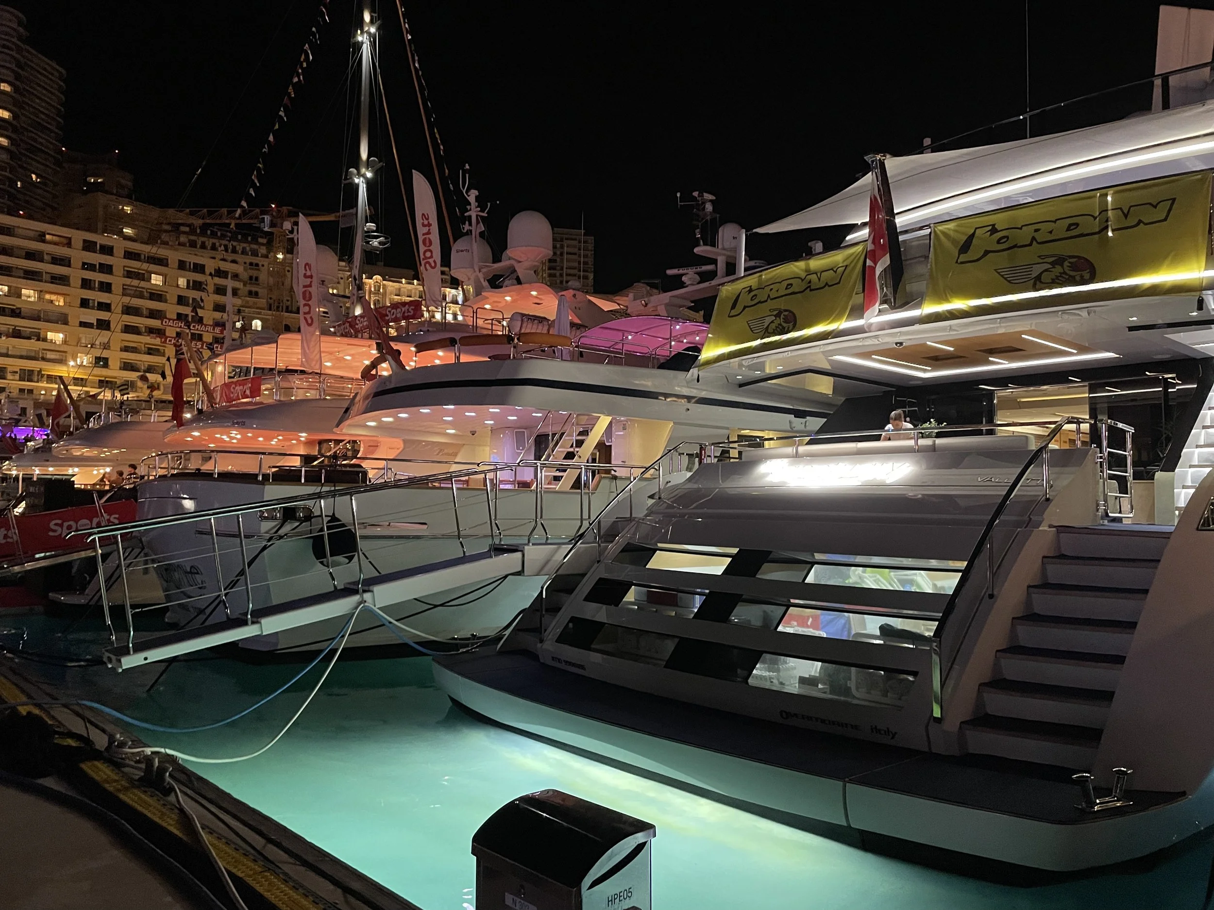 Nighttime scene at a marina with several illuminated luxury yachts docked side by side, with a high-rise building in the background.