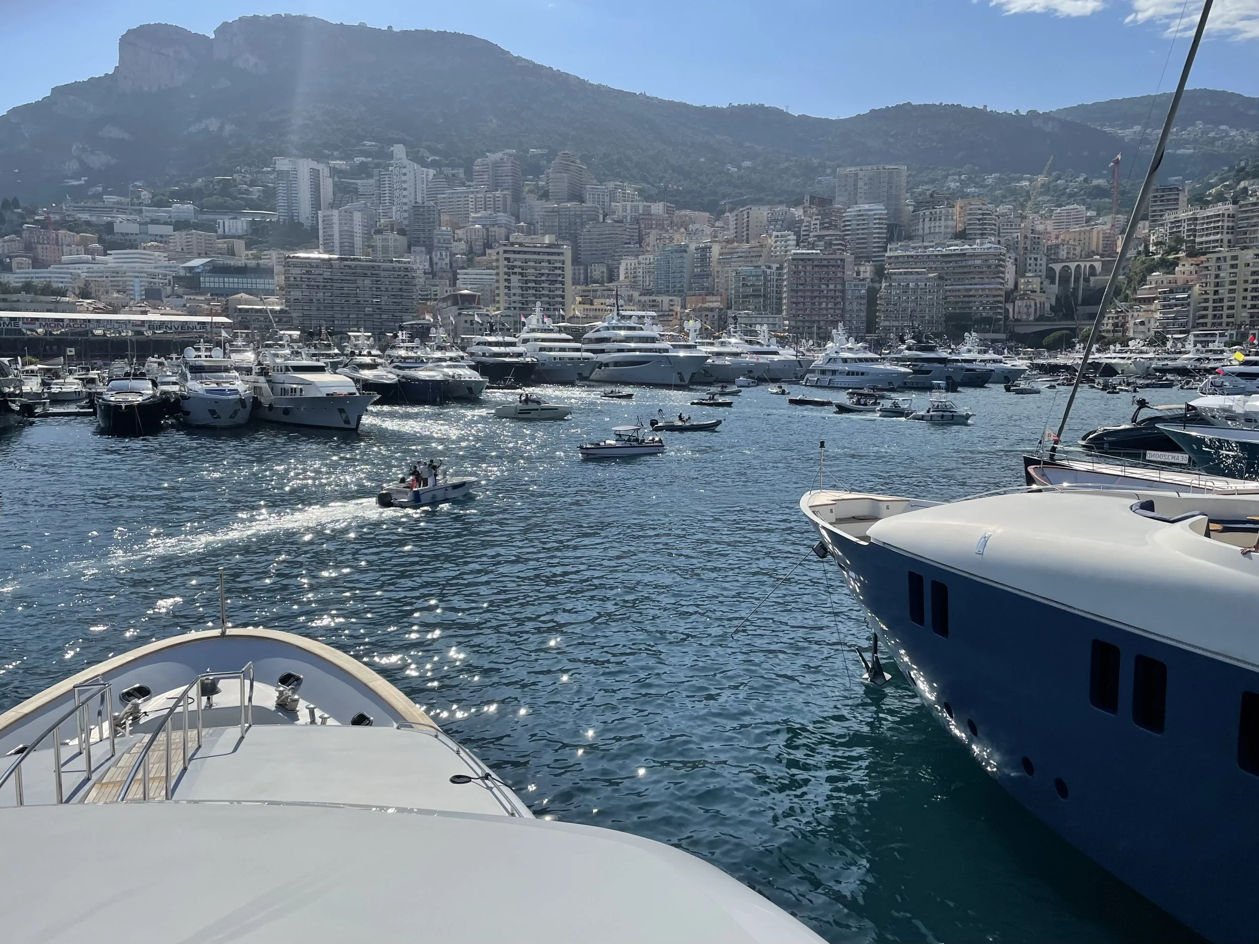 View of a marina with numerous yachts and boats docked, city buildings and hills in the background, and a small boat moving across the water.
