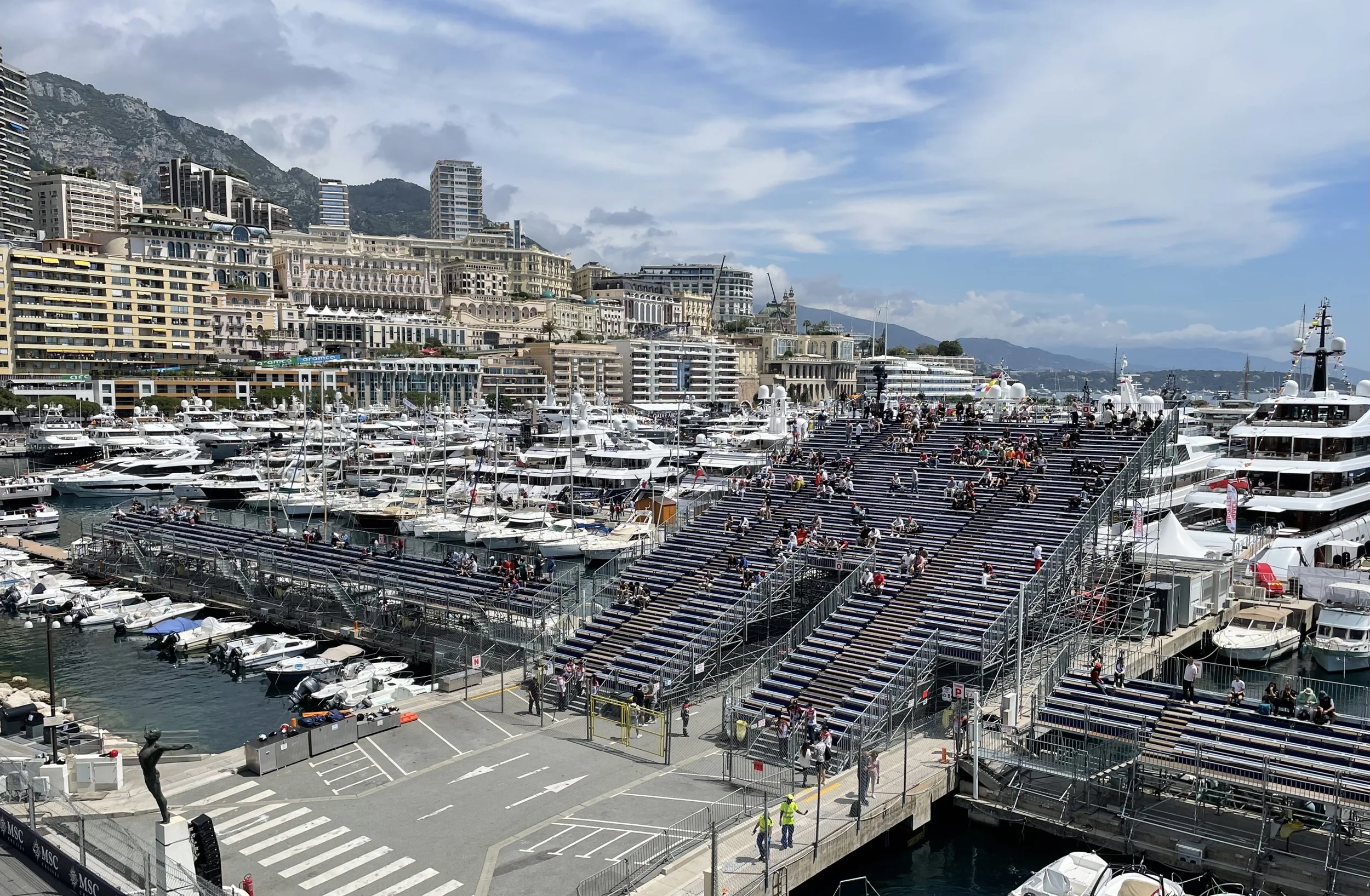 A marina filled with yachts and boats, with tiered seating and people sitting and walking. Tall buildings and mountains are visible in the background under a partly cloudy sky.