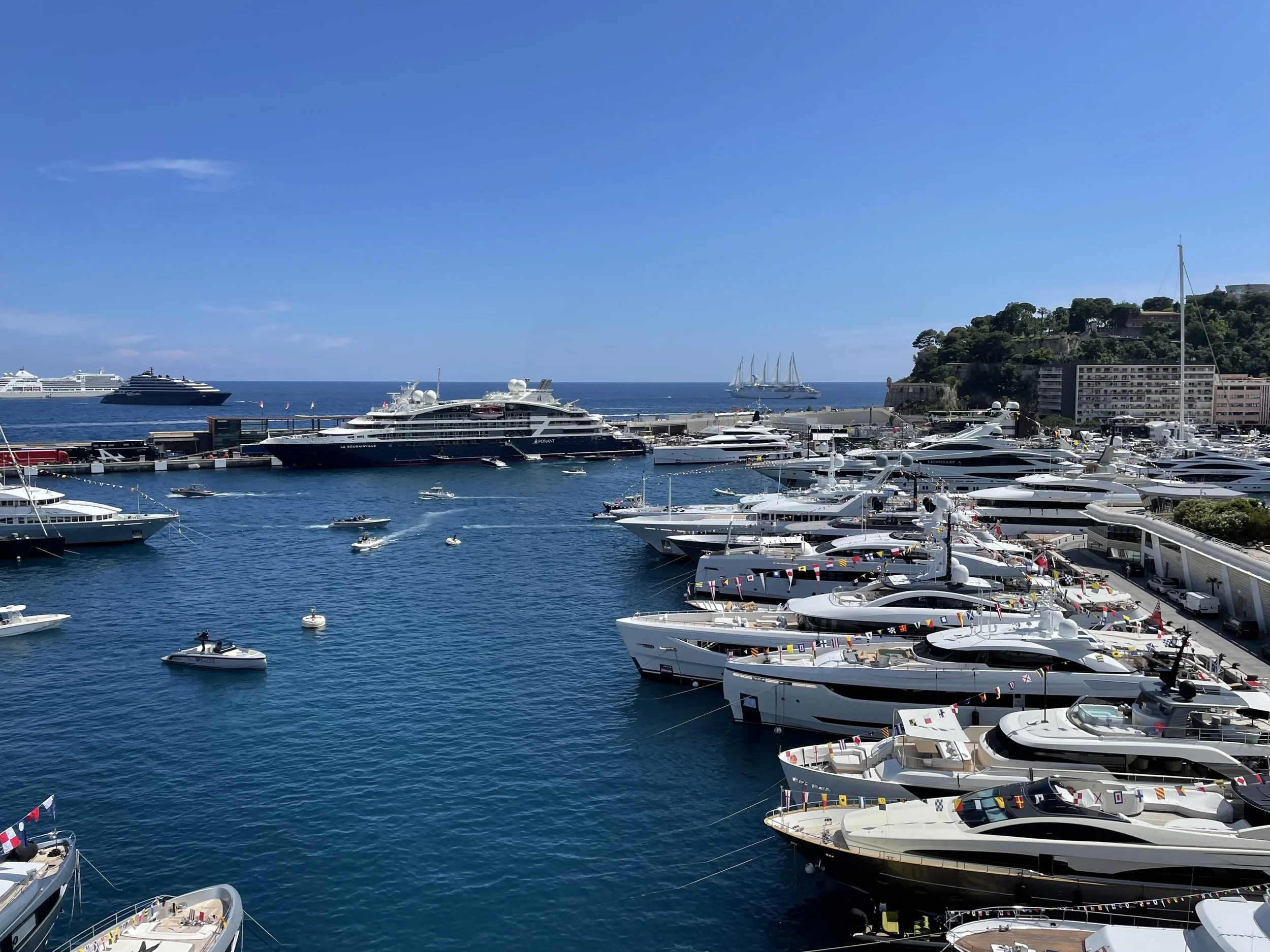 View of a marina filled with luxury yachts and boats under a clear blue sky, with buildings and a hill in the background.