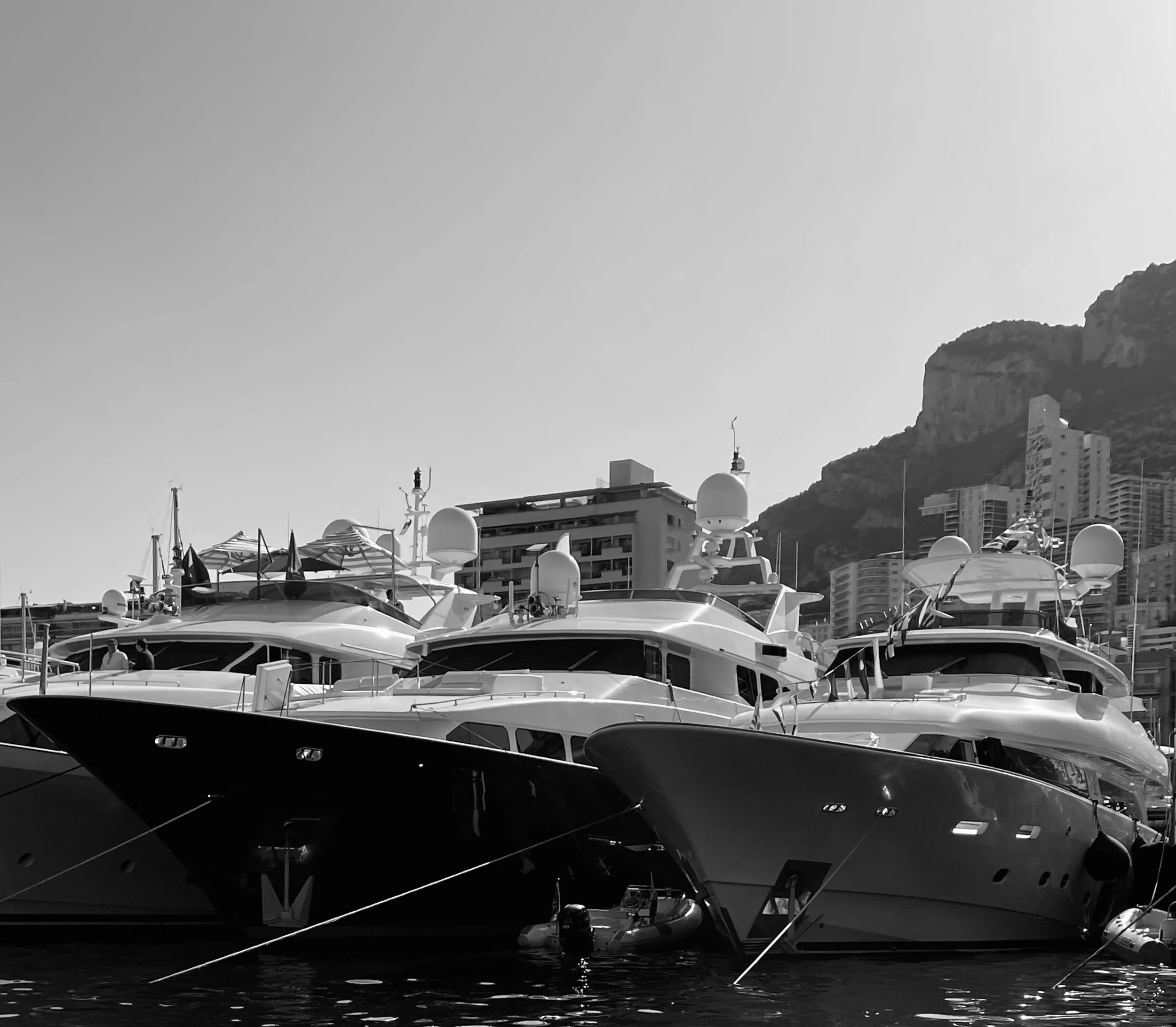Black and white photo of several luxury yachts docked at a marina with buildings and a mountain in the background.