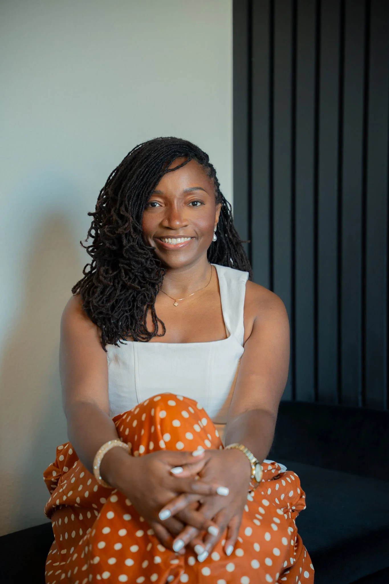 Mbu, financial coach and founder of The Audacity Way, smiling in a white top and orange polka dot skirt against a dark slatted panel