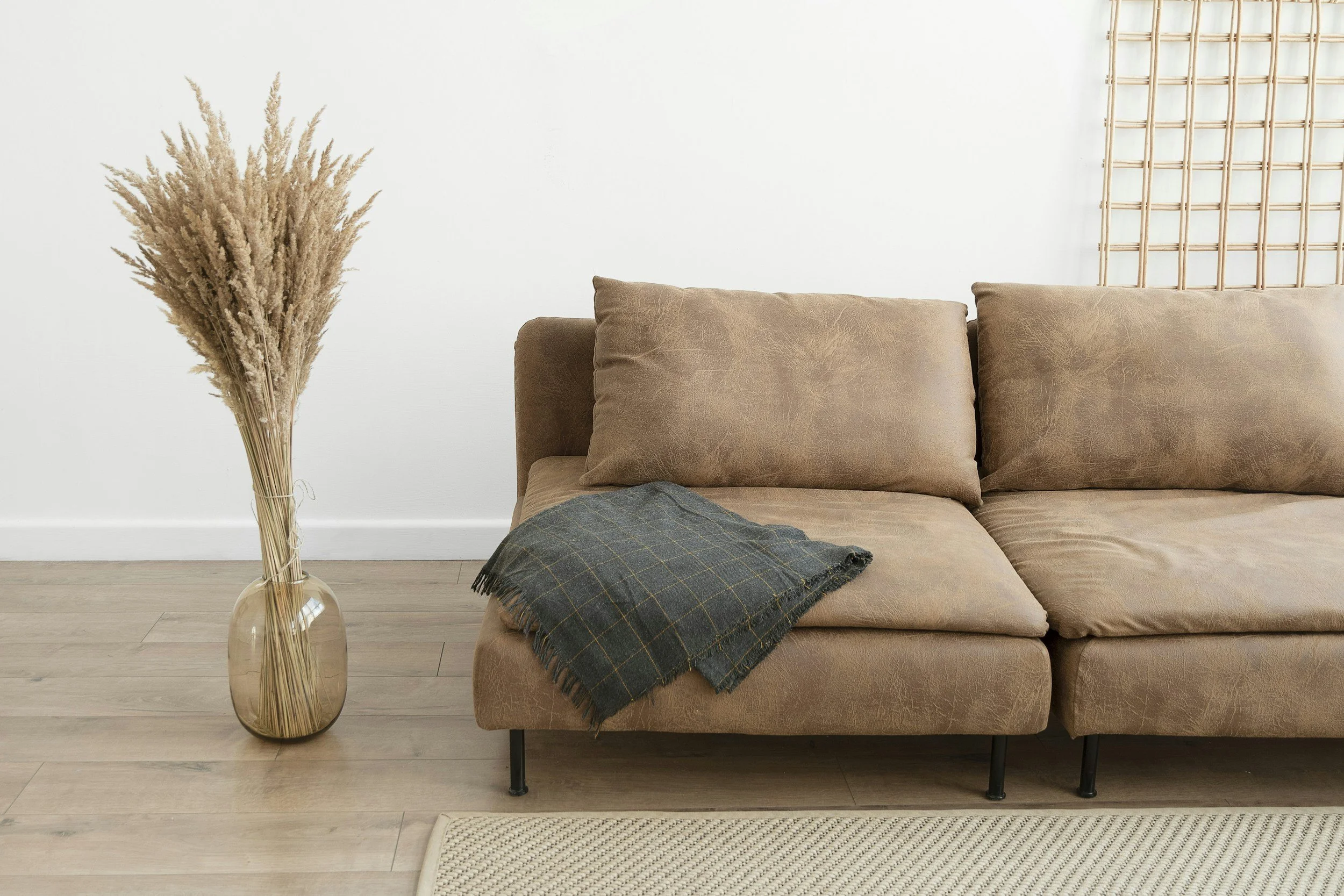 A living room with a beige leather sofa, a plaid throw blanket, a dried floral arrangement in a glass vase, a white wall, and a wooden floor.