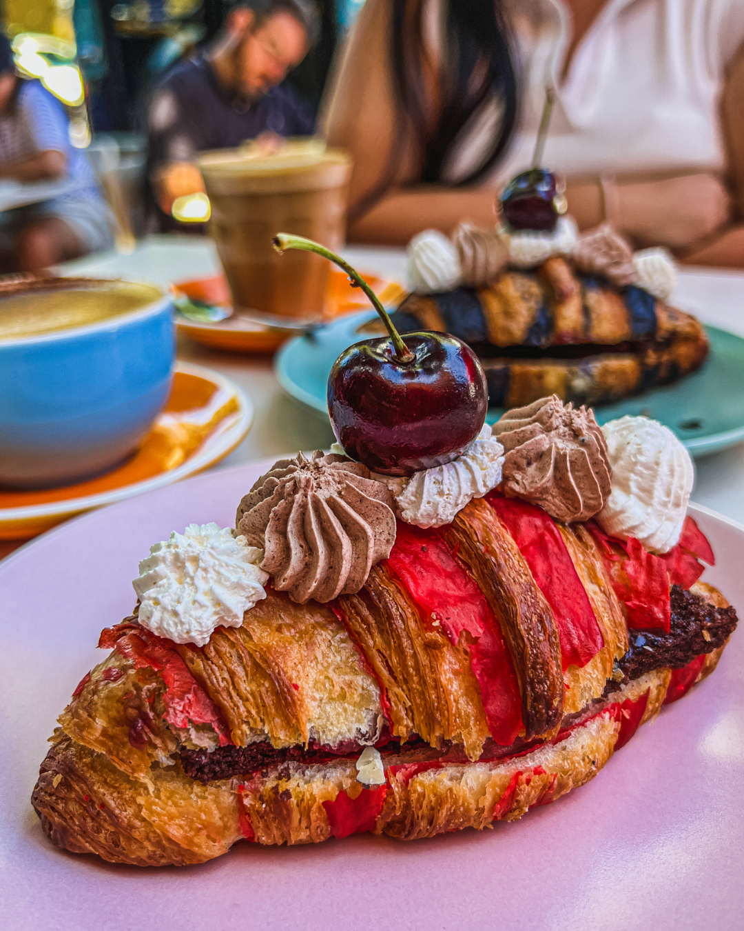 Close-up of a cherry-topped pastry with whipped cream, chocolate, and white frosting, on a pink plate, with cups of coffee and another pastry in the blurred background.