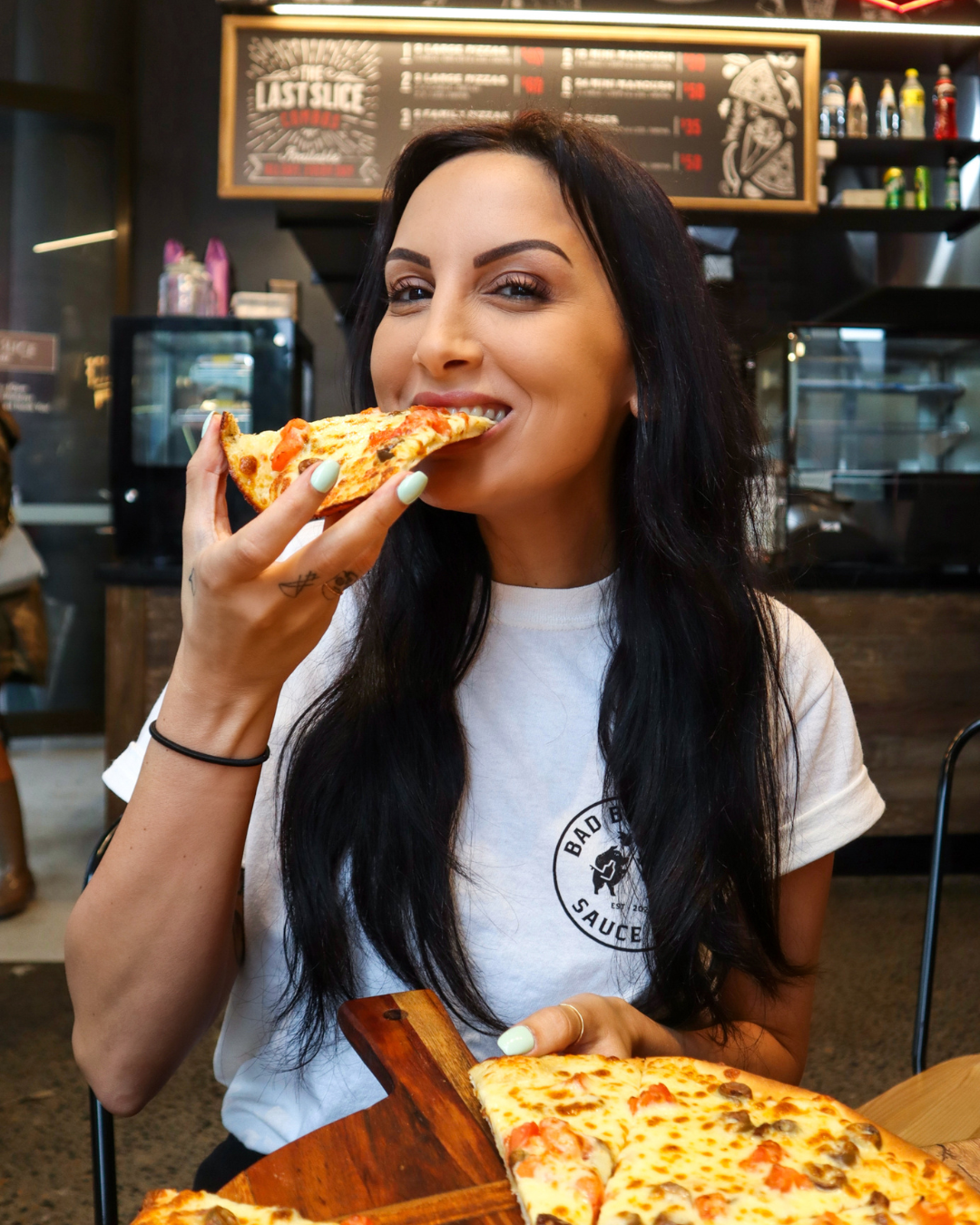 A woman with long black hair and light green nails eating a slice of pizza in a pizzeria restaurant.