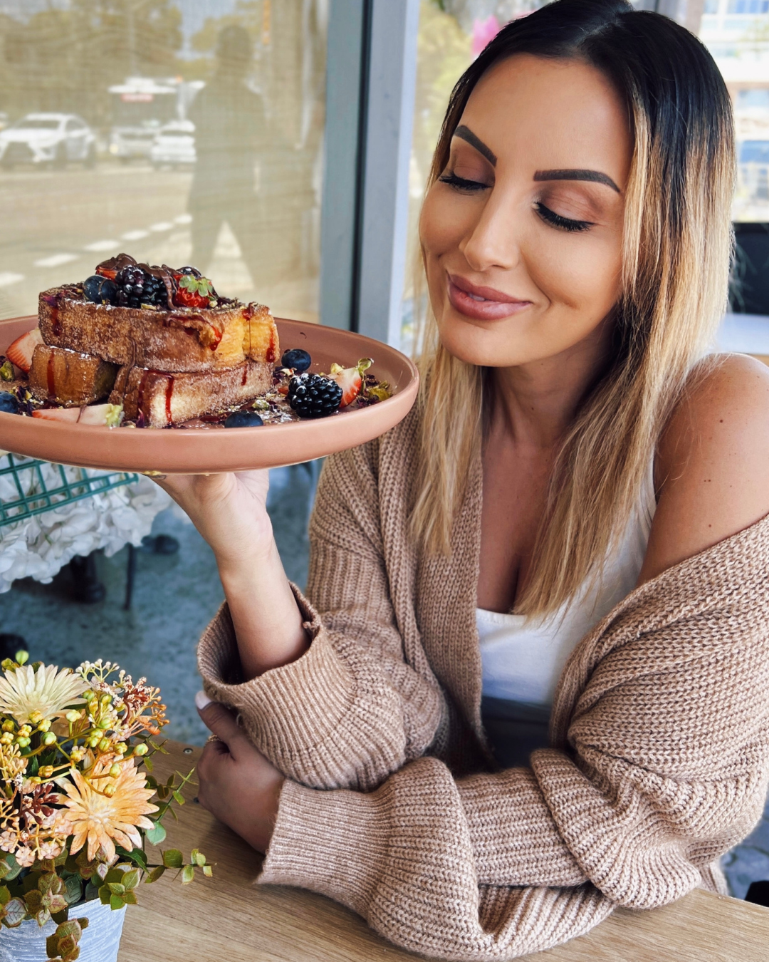 A woman holding a pink plate with French toast topped with fresh berries, syrup, and powdered sugar, smiling with her eyes closed.