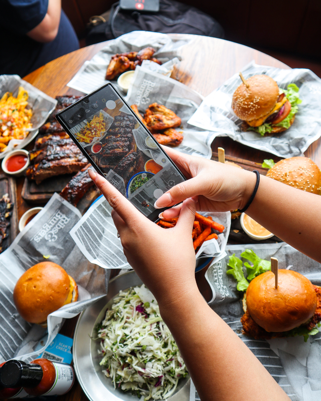 A person taking a photo of a spread of barbecue food and burgers on a wooden table.