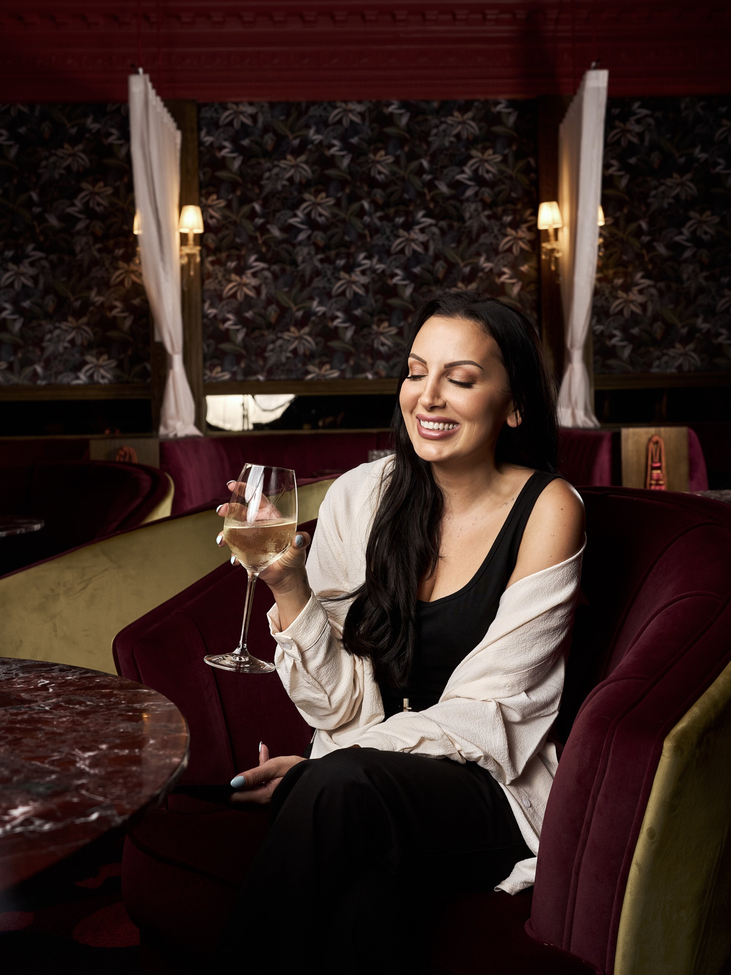 A woman with long dark hair is smiling and holding a glass of white wine in a restaurant with vintage wallpaper and soft lighting.