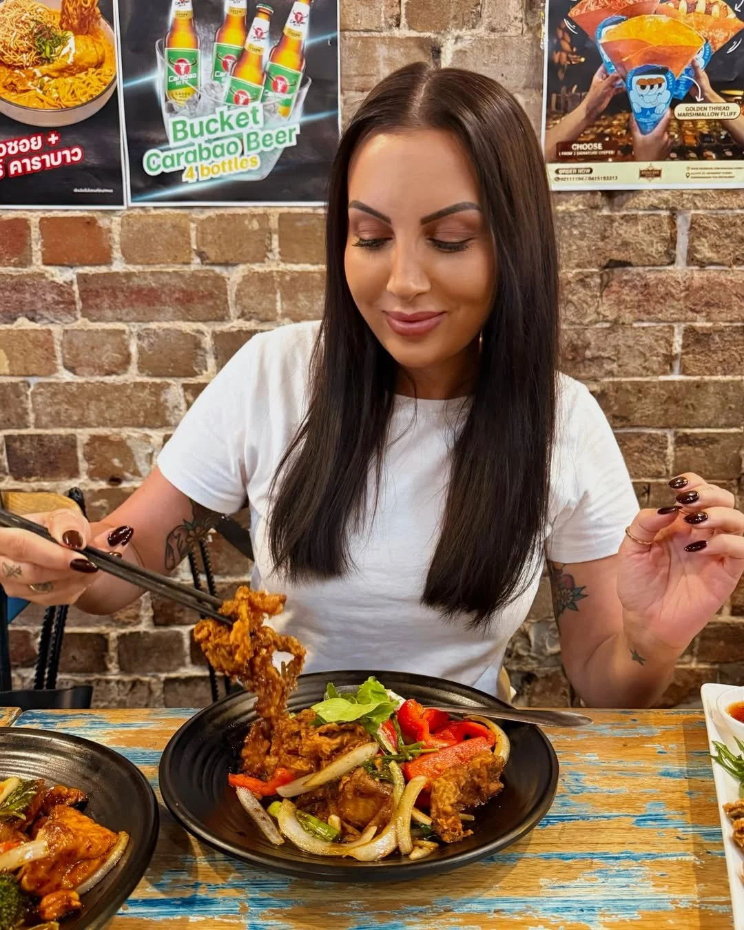 A woman with long dark hair and tattoos on her arms is eating a plate of fried chicken and vegetables at a restaurant with a brick wall background. There are promotional posters on the wall.