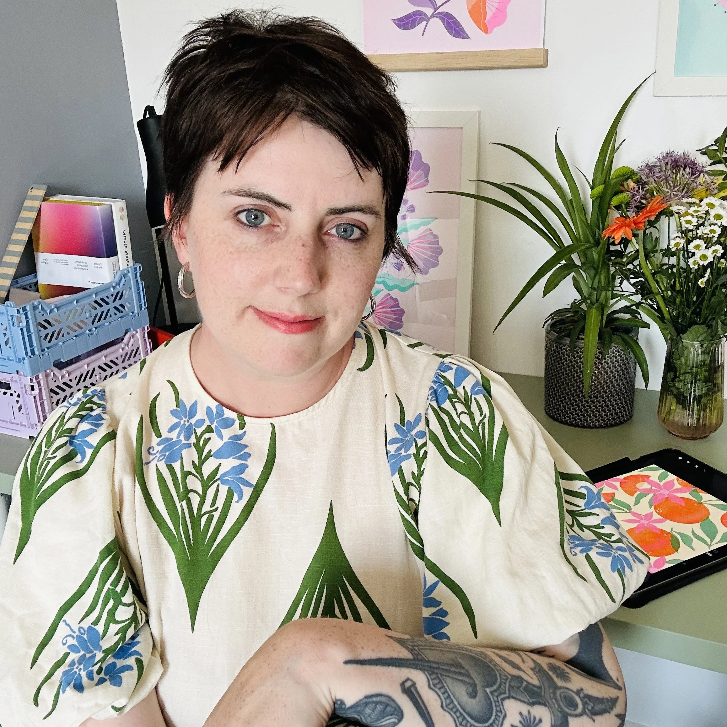 A woman with short dark hair and blue eyes, wearing a cream-colored shirt with a green and blue floral pattern, sitting at a desk with colourful flowers and stationery around her.