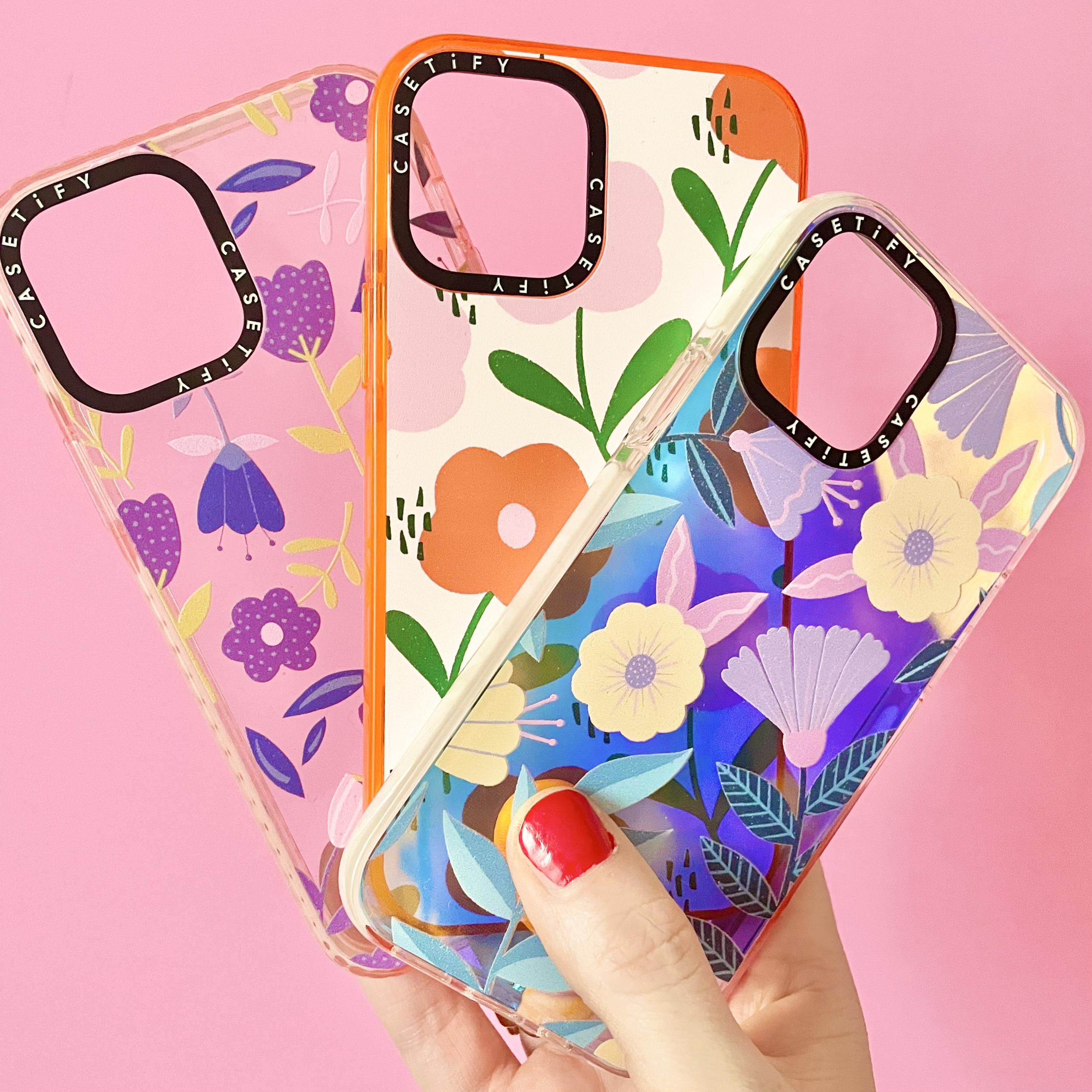 Three colourful phone cases with floral patterns, held by a person with a red-painted thumbnail, on a pink background.
