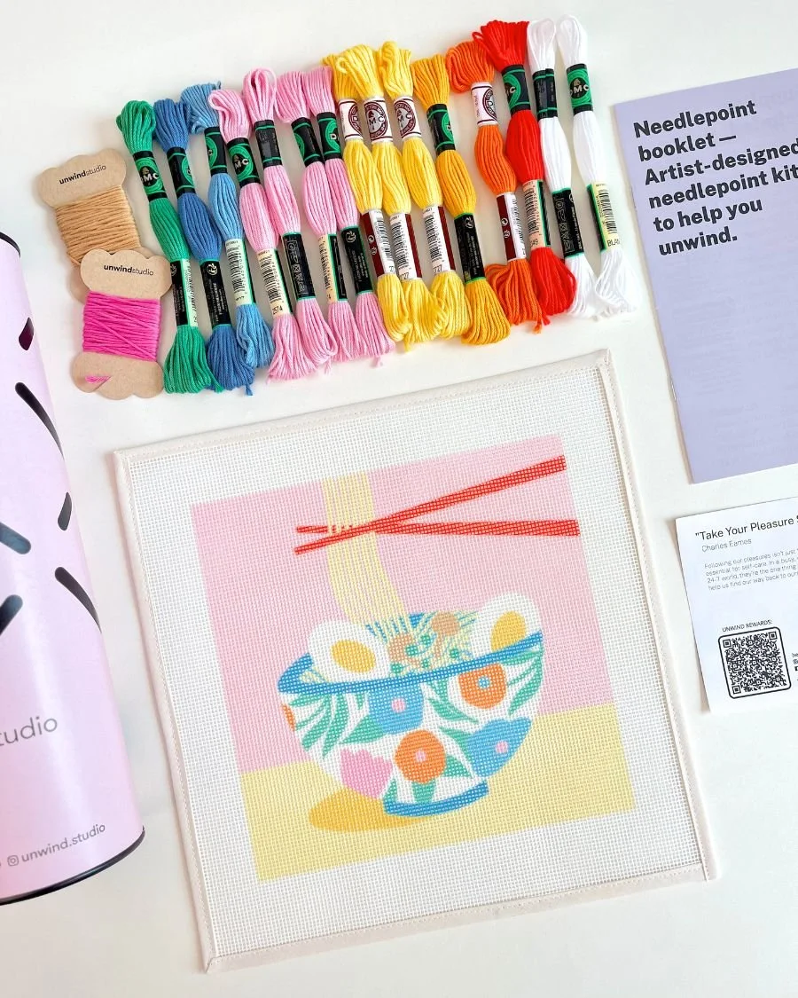 Colorful embroidery floss, including beige, pink, green, blue, yellow, orange, and white, arranged on a table. Next to the floss is a printed needlepoint kit with a colorful bowl of fruit design, and a purple informational sheet about needlepoint kit