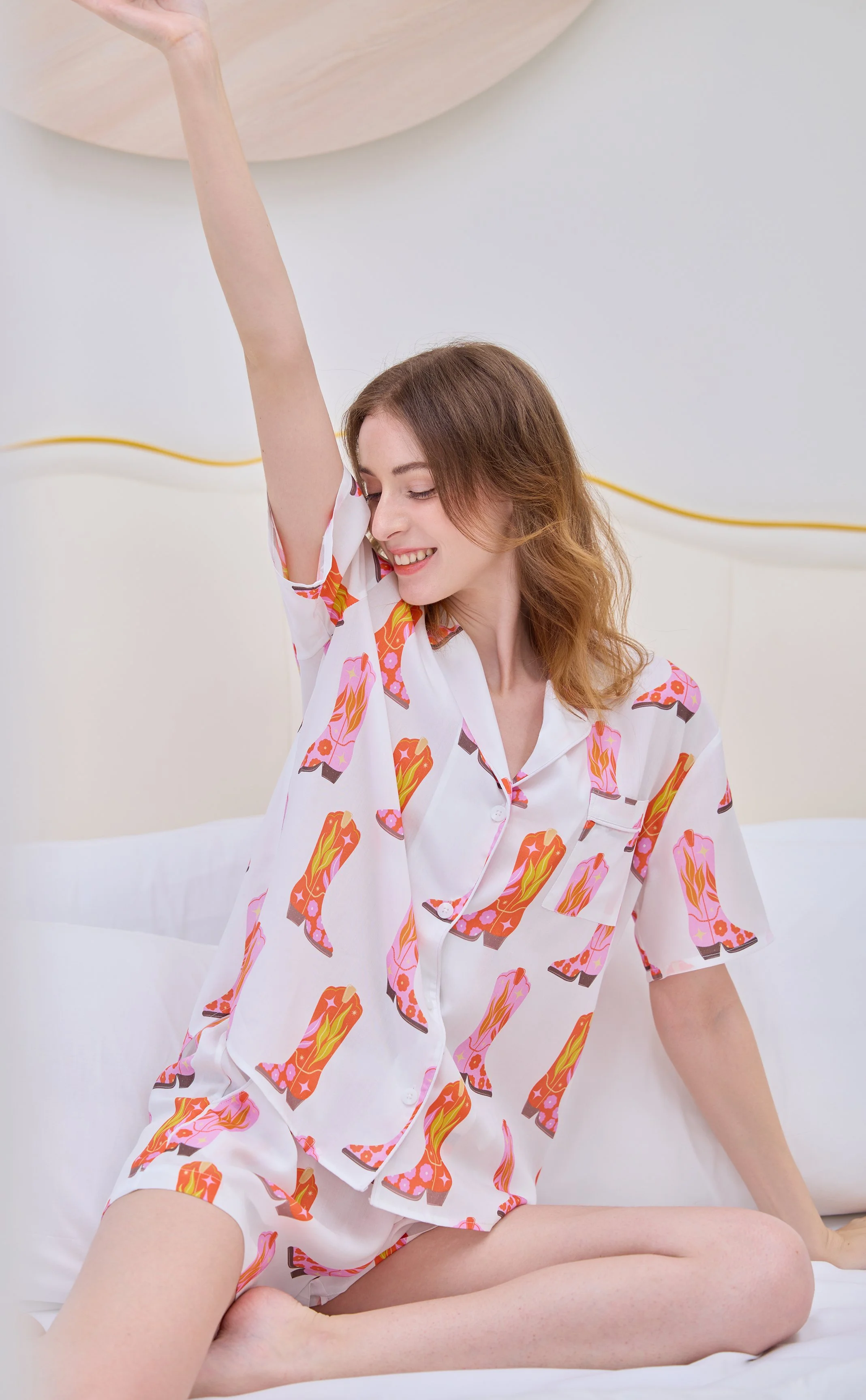 Young woman with ginger hair, wearing pajama top and shorts with a cowboy boot pattern, sitting on a bed and stretching her arm upward with a smile.