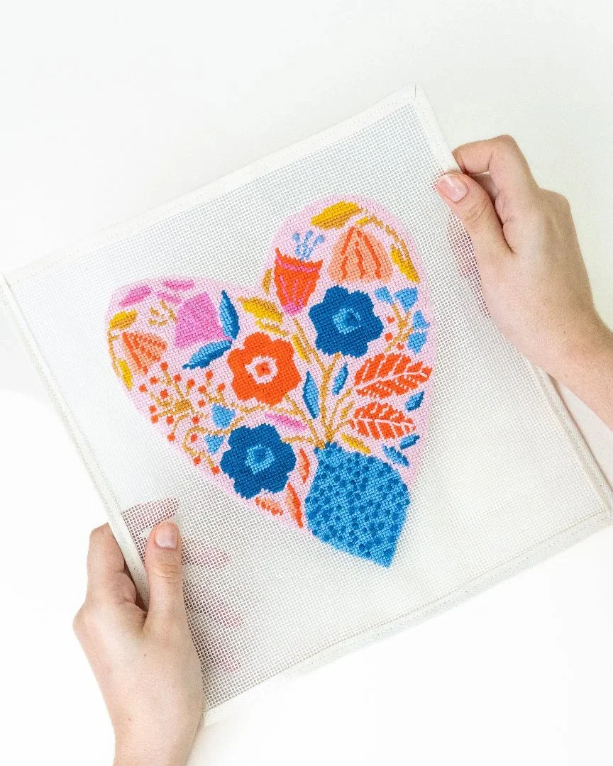 Cross-stitch embroidery of a heart made up of colourful flowers on white fabric.