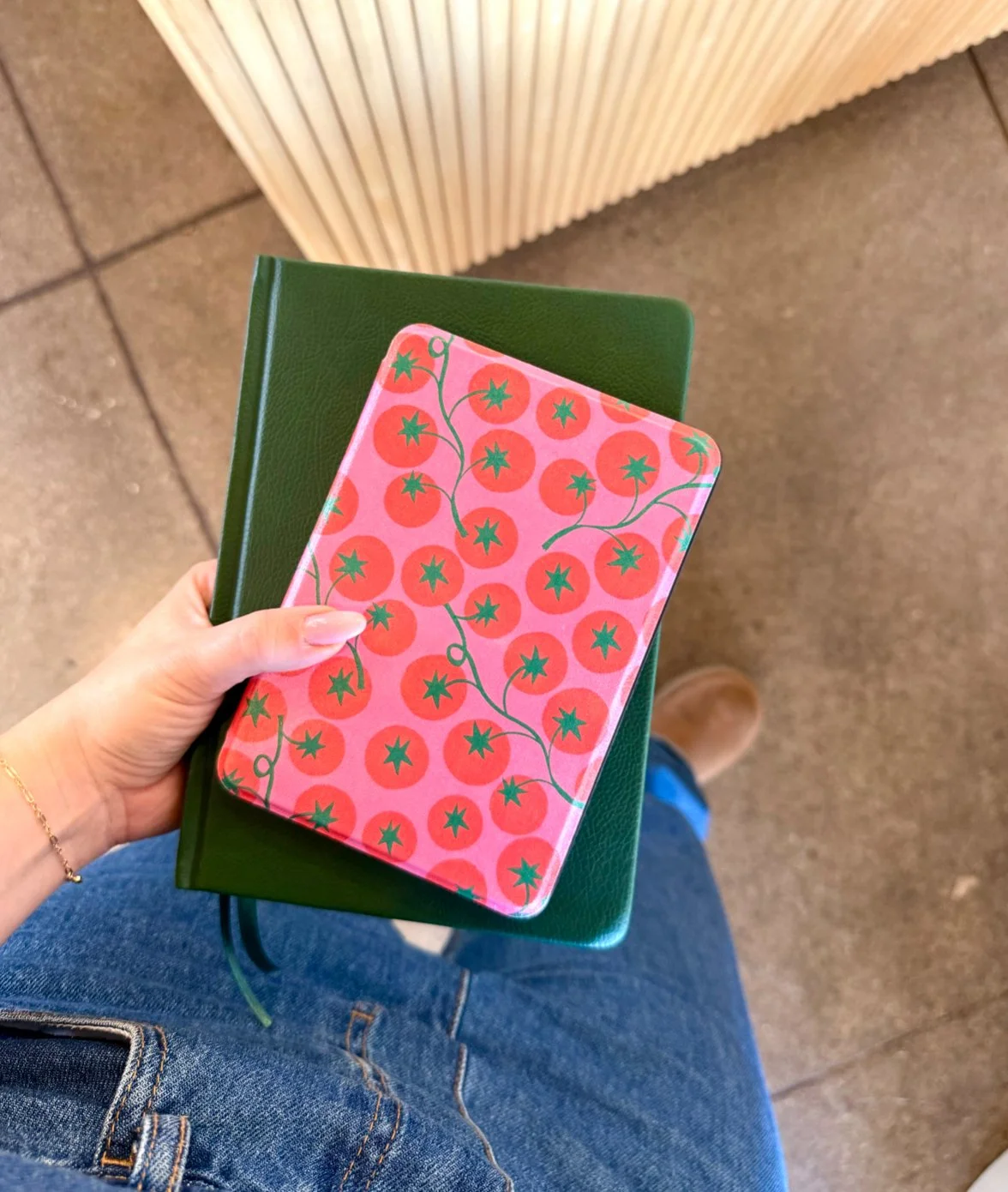 Hand holding a pink notebook with a tomato pattern, resting on a closed green notebook, above denim jeans and a brown shoe, on a beige tiled floor.