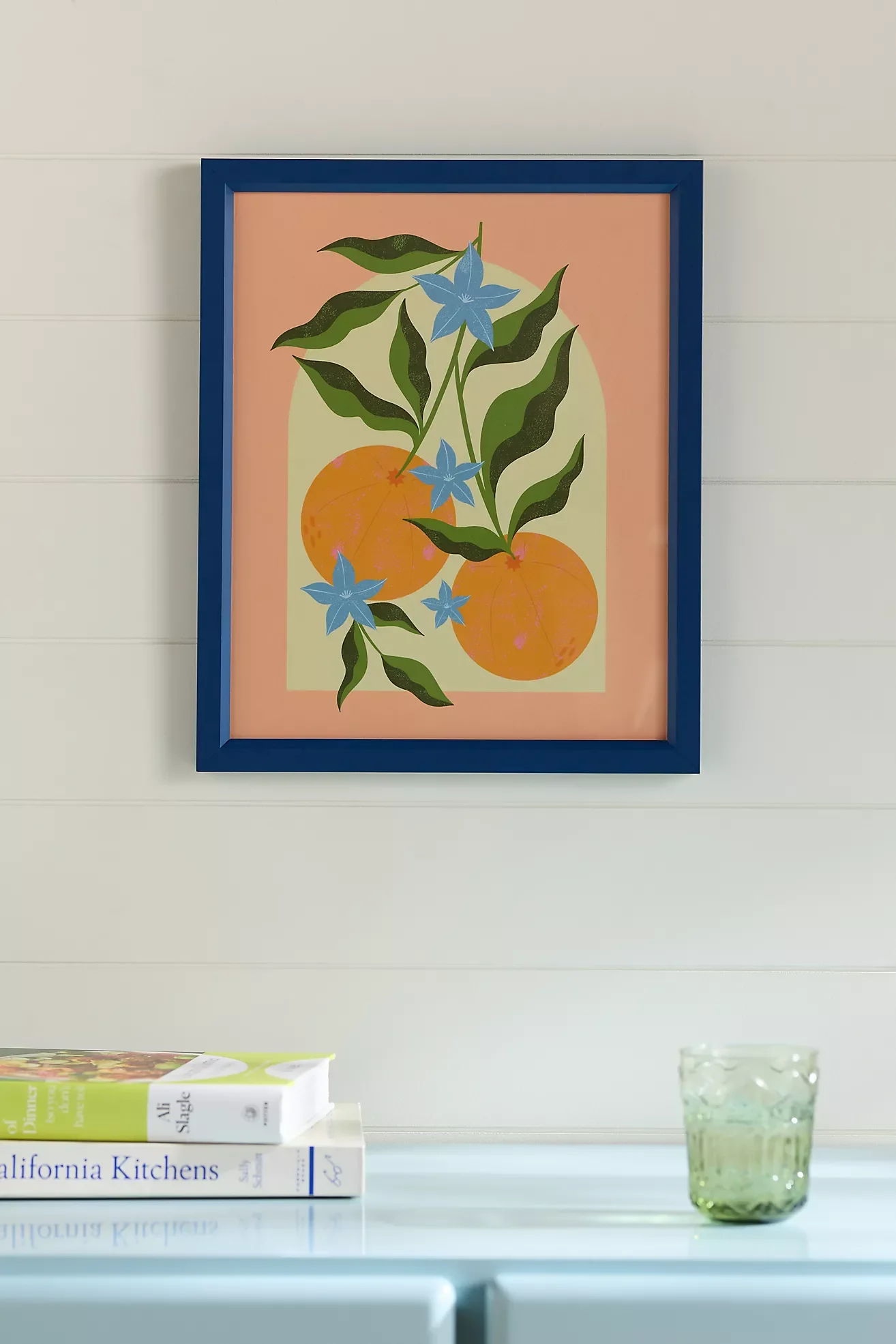 Framed botanical art print of oranges, green leaves, and blue flowers on a peach and cream background, hanging on a wall above books and a glass.