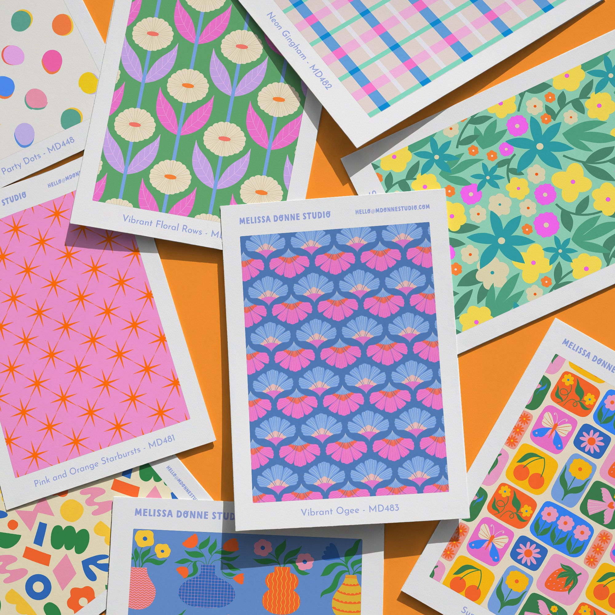 A collection of colourful floral and geometric pattern designs on paper sheets, laid out on an orange surface. Each sheet features bright, vibrant illustrations of flowers, leaves, and shapes.