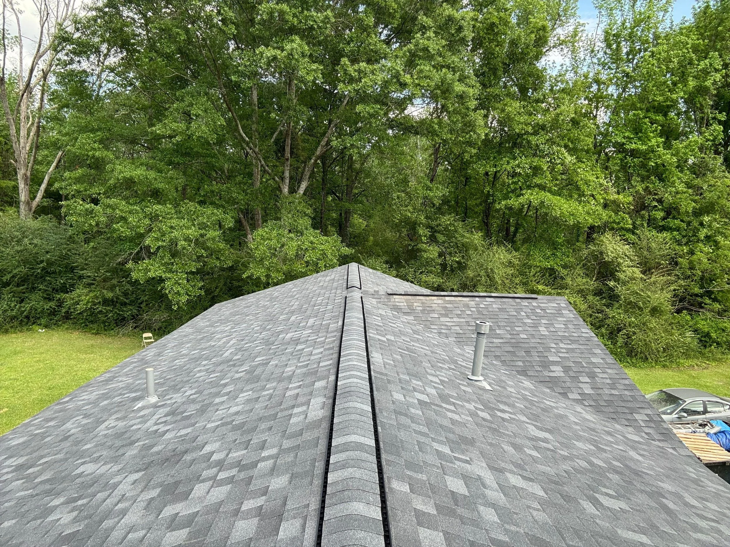 View of a house roof with gray shingles, two vent pipes, and a black cable or wire running down the center. In the background, there is lush green trees and a partial view of a car and yard.