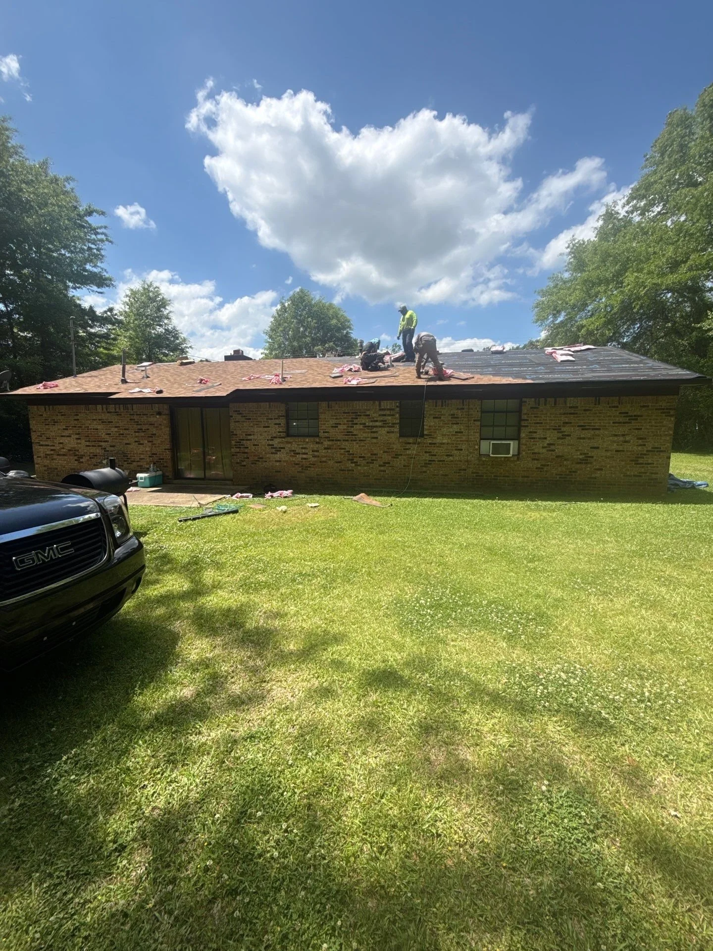A house with a roof under repair on a sunny day with a blue sky and clouds. Several workers are on the roof working on the shingles, with tools and materials scattered around.