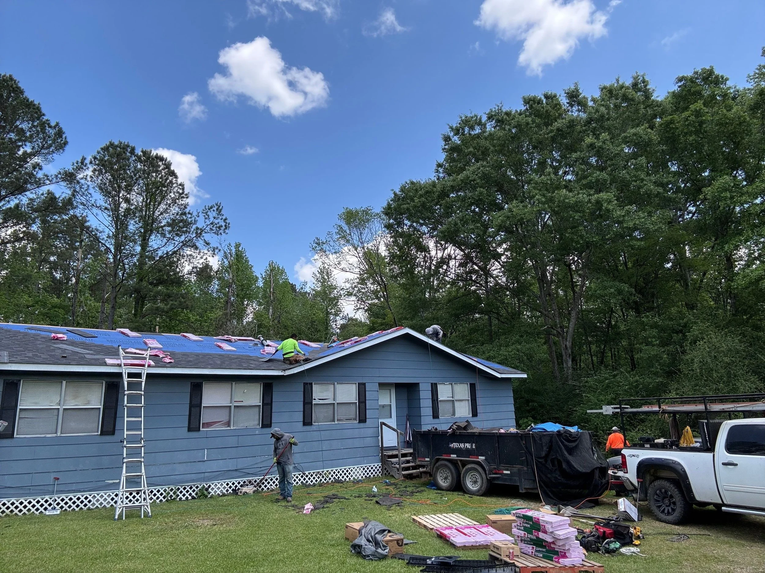 Workers installing or repairing a blue house roof with shingles, tools, and materials in a backyard with trees and a clear sky.