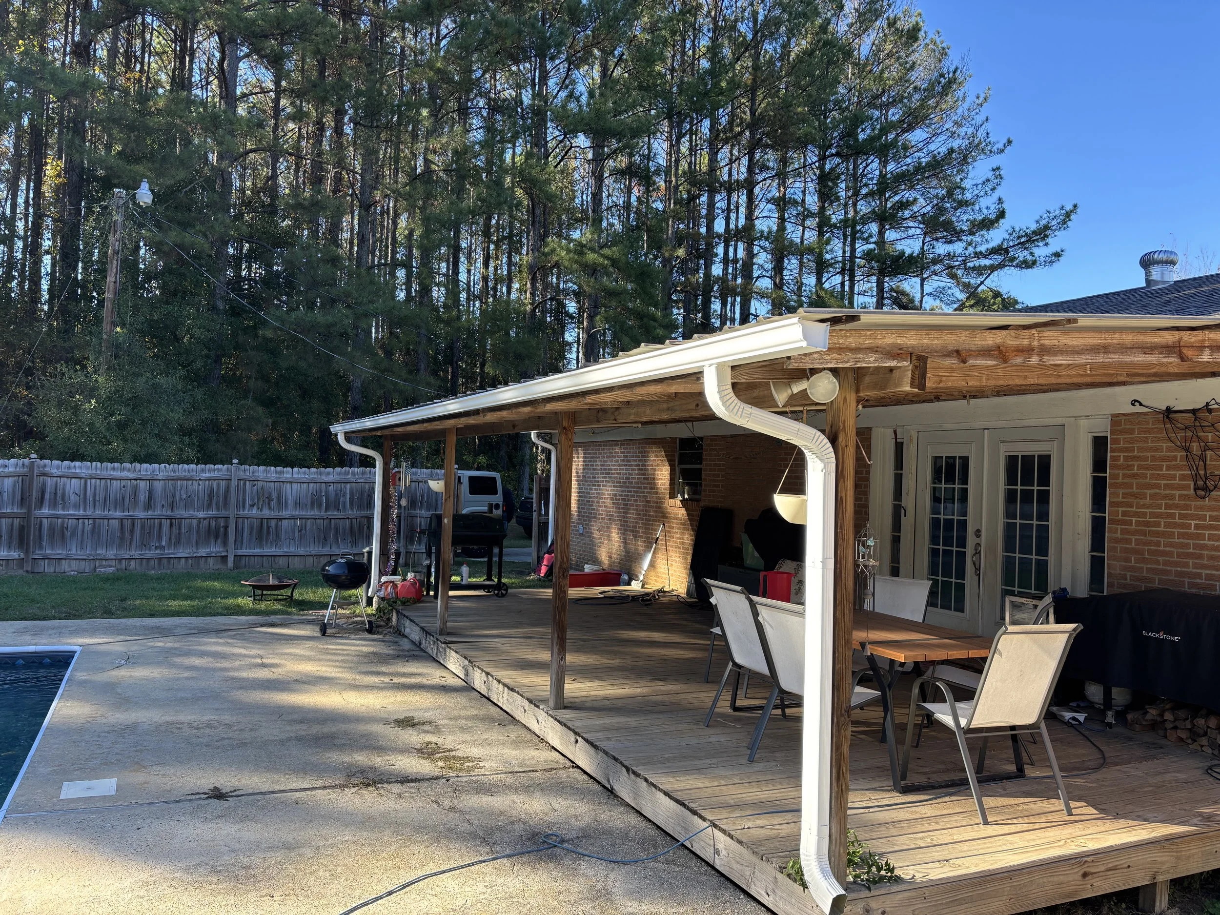 Backyard patio with a wooden deck, outdoor furniture, a barbecue grill, and a covered area with a roof, surrounded by a wooden fence and tall trees, with a swimming pool in the foreground.