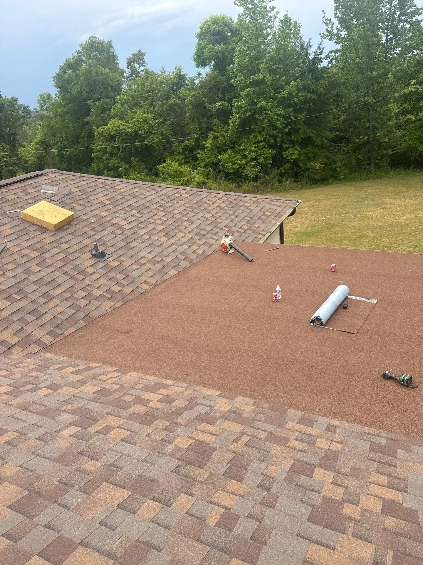 A rooftop with shingles in the process of being replaced or repaired. Tools, a roll of roofing material, and cans are scattered on the roof. Green trees and a cloudy sky are in the background.