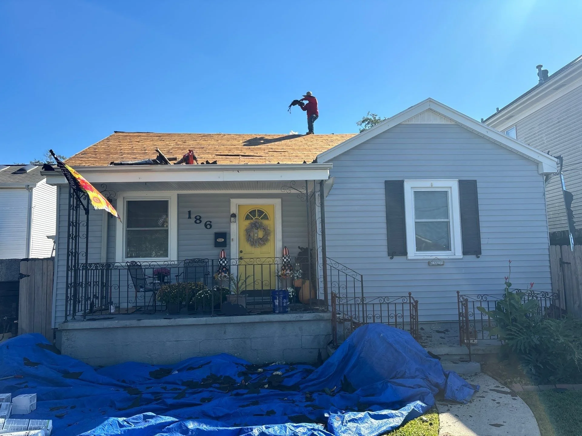 A person on a roof appears to be replacing or repairing shingles. The house in front of the roof is a white, single-family home with a decorated front door and flowers on the porch. There is blue tarp on the ground in front of the house, likely for c
